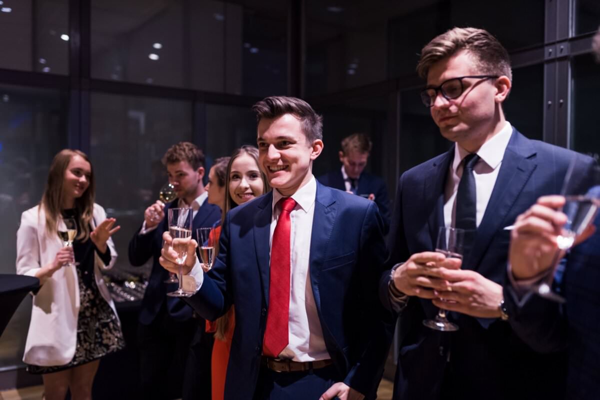 Eine Gruppe formell gekleideter Menschen hat sich in einem Raum versammelt und trinkt Gläser mit Champagner. Sie sehen aus, als befänden sie sich auf einem gesellschaftlichen Ereignis oder einer Feier, wunderschön eingefangen von einem Eventfotografen Warschau. Im Vordergrund stehen zwei Männer in Anzug und Krawatte, die lächeln und sich mit anderen unterhalten.