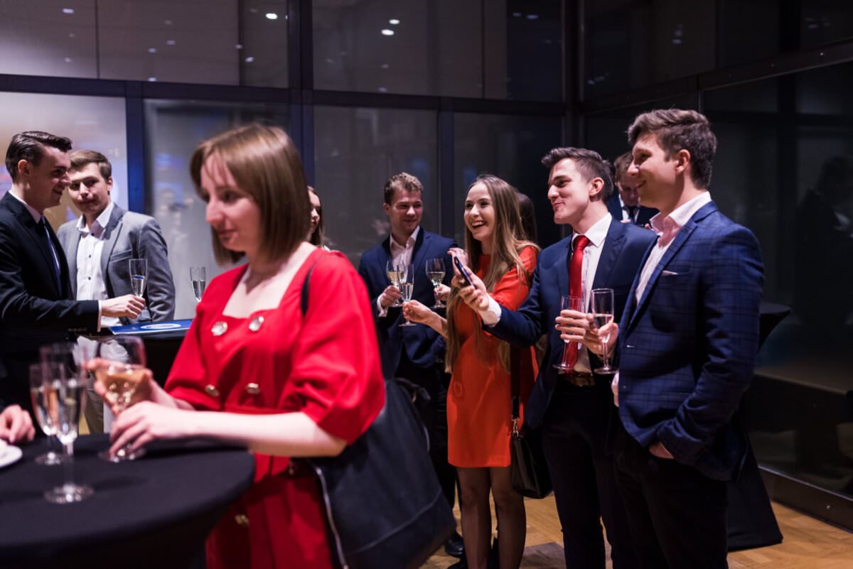 In einem modernen Innenraum versammelt sich eine Gruppe förmlich gekleideter Menschen, die Getränke in der Hand halten und in ein Gespräch vertieft sind. Eine Frau in einem roten Kleid steht im Vordergrund, während andere lächeln und sich im Hintergrund unterhalten. Das deutet auf ein gesellschaftliches Ereignis oder eine Netzwerkveranstaltung hin - was die Eventfotografie perfekt einfängt.
