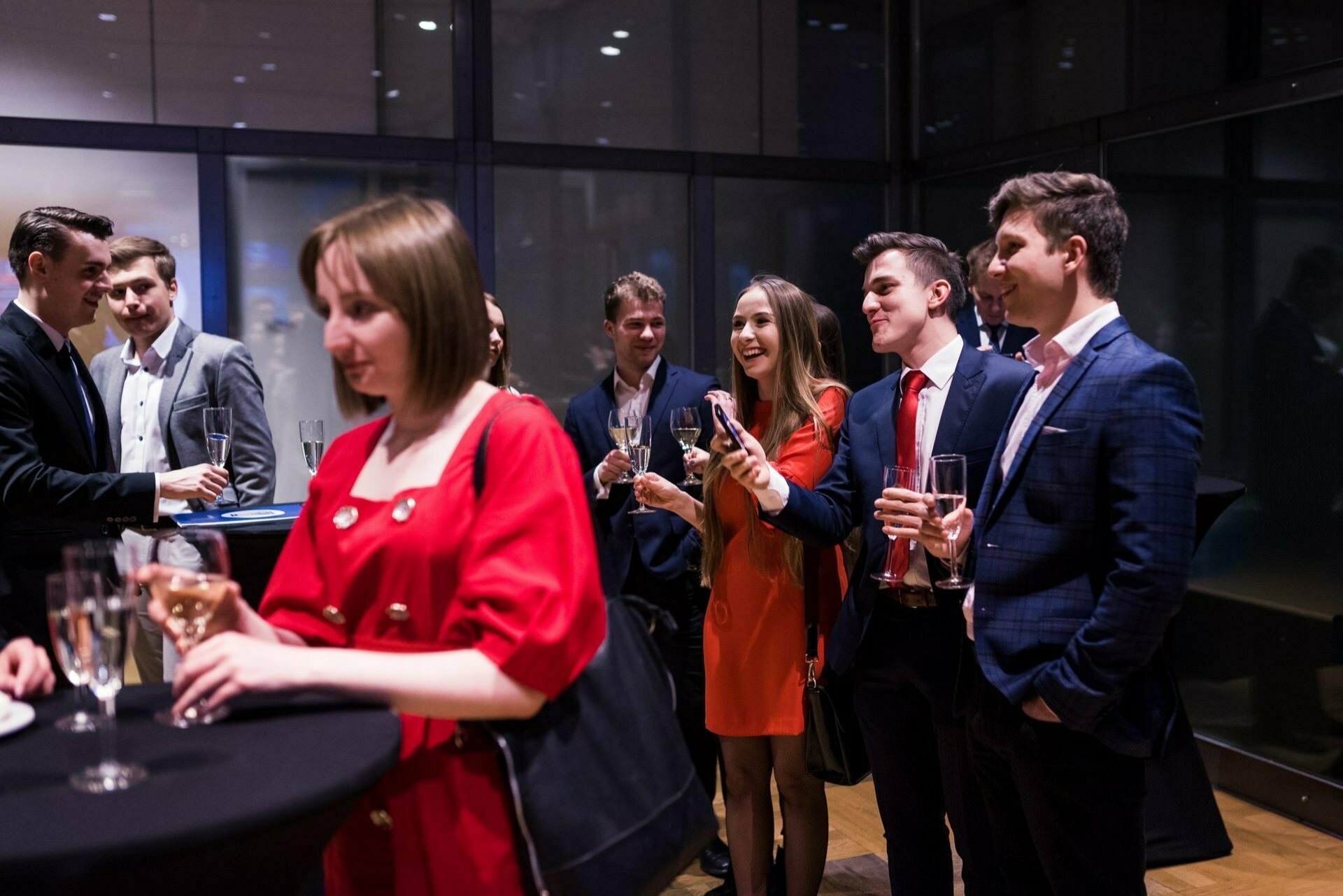 In a modern indoor space, a group of formally dressed people gather, holding drinks and immersed in conversation. One woman in a red dress is in the foreground, while others are smiling and interacting in the background, suggesting a social or networking event - something captured perfectly by the event photography. 