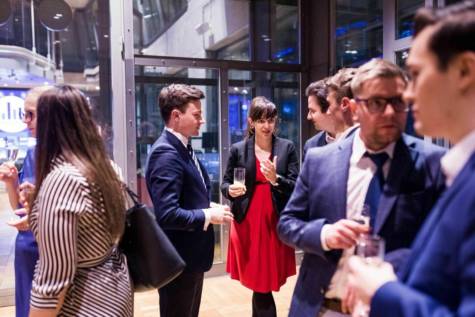 A group of people in business attire talk to each other during a networking event in a room. Some hold drinks, while a woman in a red dress carries on a conversation with a man in a suit. The room has large windows and a modern interior design, perfectly captured by event photographer Warsaw.  