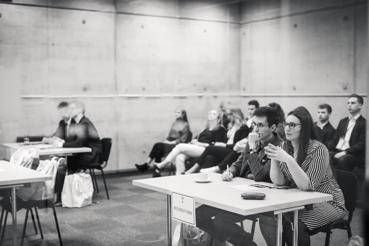 Das Schwarz-Weiß-Bild zeigt eine Gruppe von Personen, die in einem Klassenzimmer sitzen. Im Vordergrund sitzen ein Mann und eine Frau an einem Tisch mit der Aufschrift 'PRESYDIUM' und führen ein Gespräch. Hinter ihnen sitzen mehrere Personen in Reihen, die aufmerksam zuhören und die Essenz der Eventfotografie einfangen.