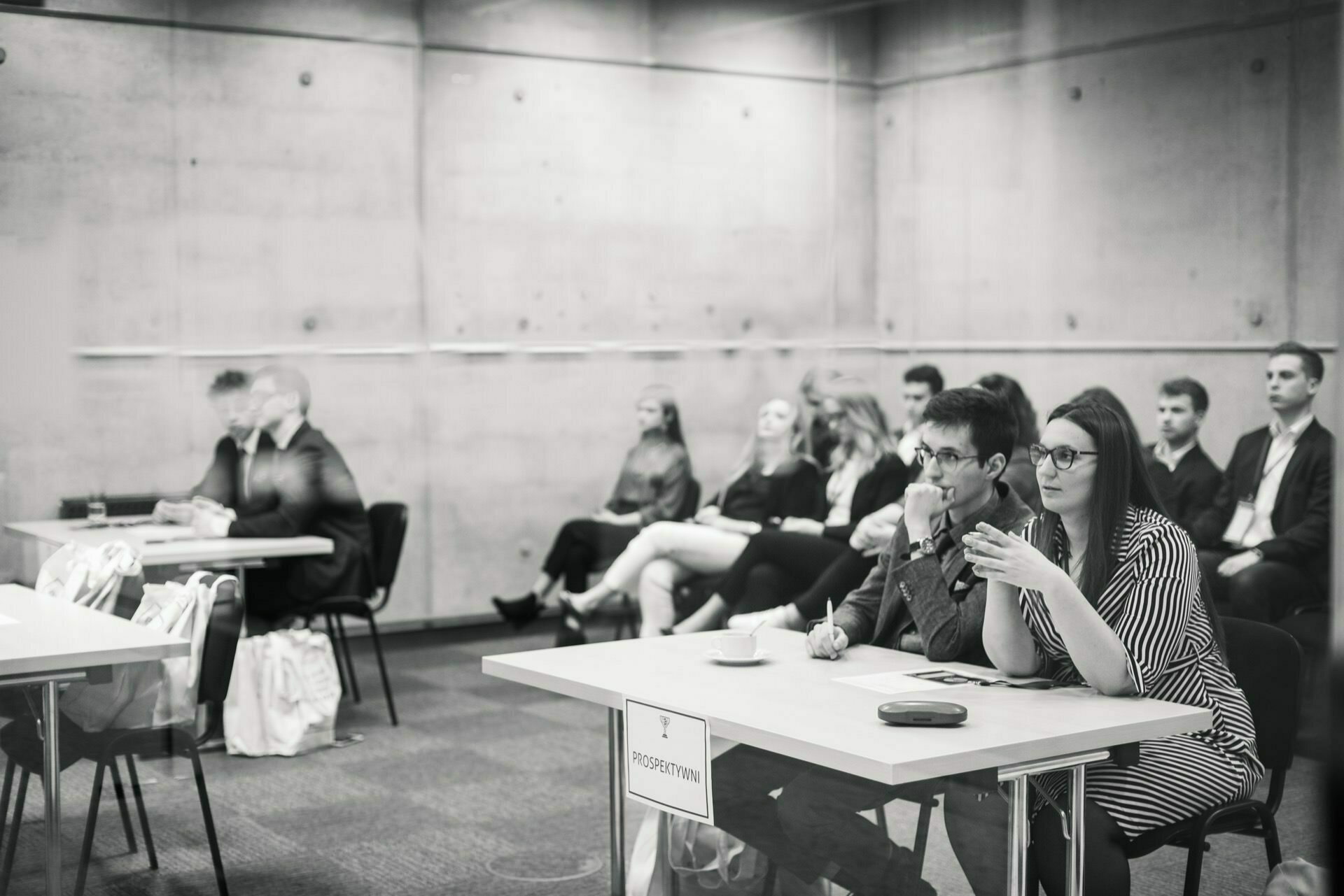 The black and white image shows a group of people sitting in a classroom. In the foreground, a man and a woman sit at a table labeled "PRESIDIUM" and are engaged in conversation. Behind them, several people are sitting in rows, attentively listening and capturing the essence of event photography.  