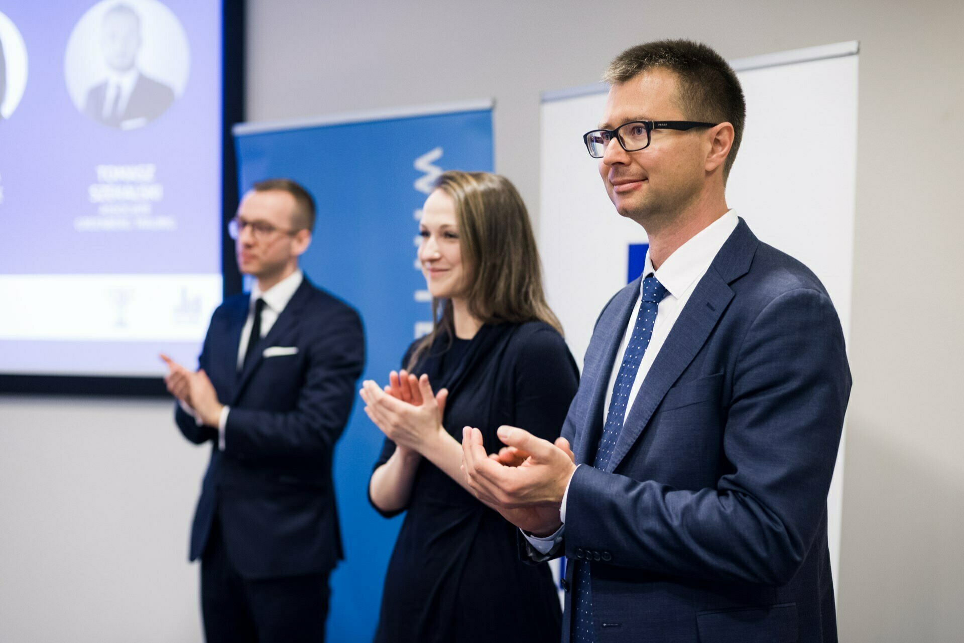 Three people dressed in business attire stand and clap at an official event. Behind them are banners and a screen displaying partial text and images. This photo essay captures the essence of a professional setting, creating excellent event photography by event photographer Warsaw.  