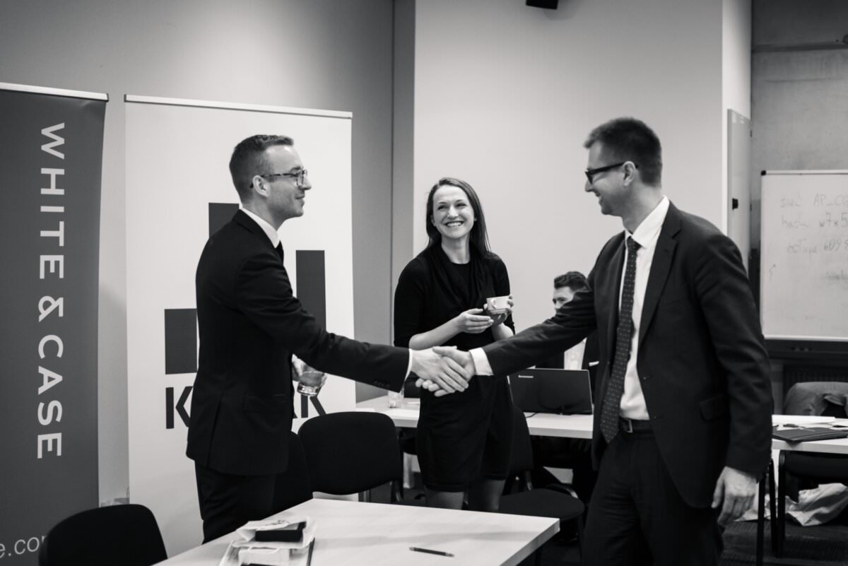 Auf diesem Schwarz-Weiß-Foto eines professionellen *Eventfotografie*-Moments befinden sich drei Personen in einem Konferenzraum. Zwei Männer in Anzügen schütteln sich die Hände, während eine Frau in einem Kleid zwischen ihnen steht, eine Tasse hält und lächelt. Im Hintergrund sind Banner mit der Aufschrift 'WHITE & CASE' und einem anderen Logo zu sehen.