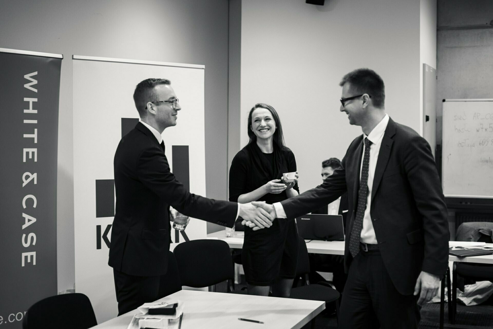 In this black and white photo depicting a professional *event photography* moment, three people are in a conference room. Two men in suits are shaking hands, and a woman in a dress stands between them, holding a cup and smiling. In the background, banners display the words "WHITE &amp; CASE" and another logo.  