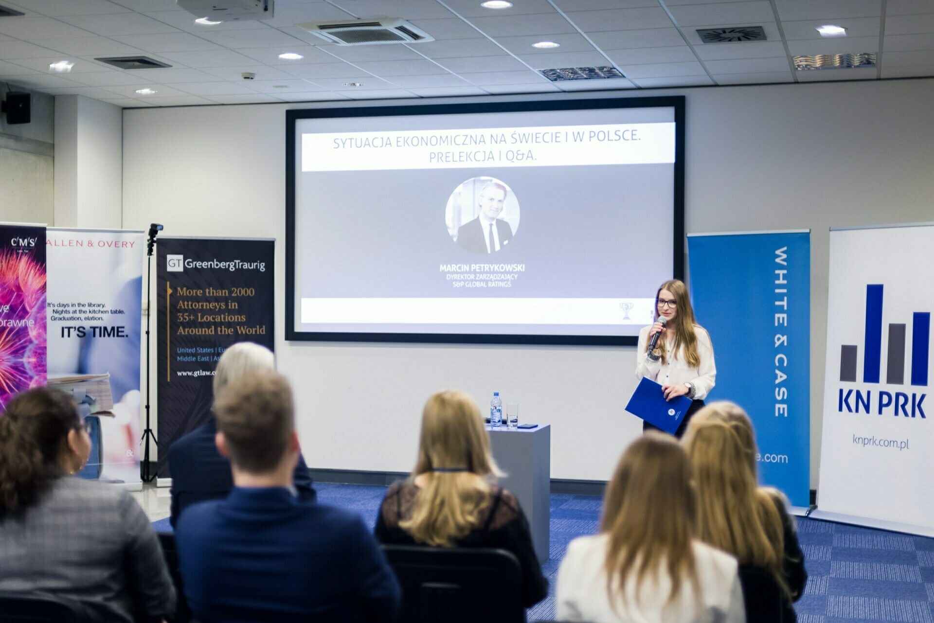 A speaker holding a microphone and a blue clipboard presents to an audience in a conference room. The slide on the screen is titled "The Economic Situation in the World And in Poland. Lecture And Q&amp;A." Seated attendees focus on the presentation, surrounded by the branding of the various sponsors - captured in an excellent photo essay of the events.   