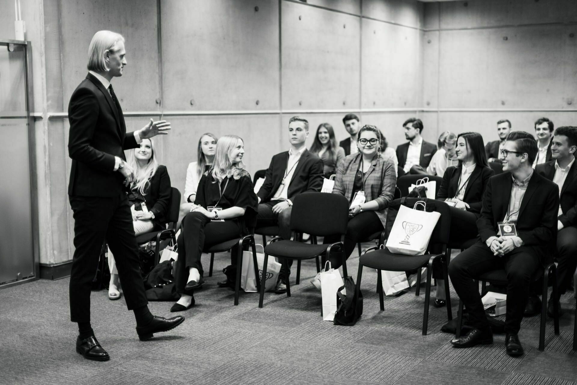 A person in a suit speaks to a room full of seated people. Participants, also dressed in formal attire, sit in rows and look engaged. Some hold notebooks or bags with a trophy symbol on them, while an event photographer Warsaw captures every moment of this modern conference.  