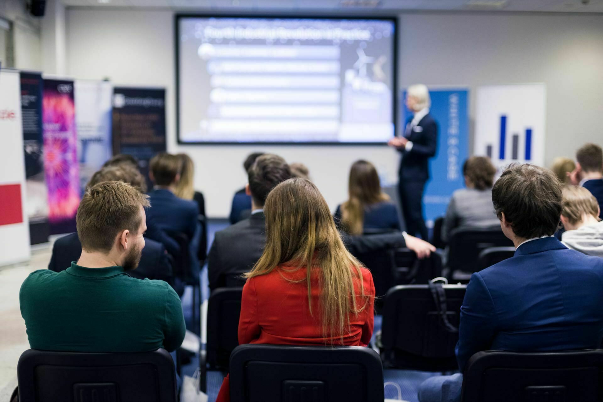 A group of people sit in a conference room in front of a presentation screen that displays blurry text and charts. A presenter in a suit stands at the front and addresses the audience. The room is decorated with various banners and posters, capturing the essence of event photography.  