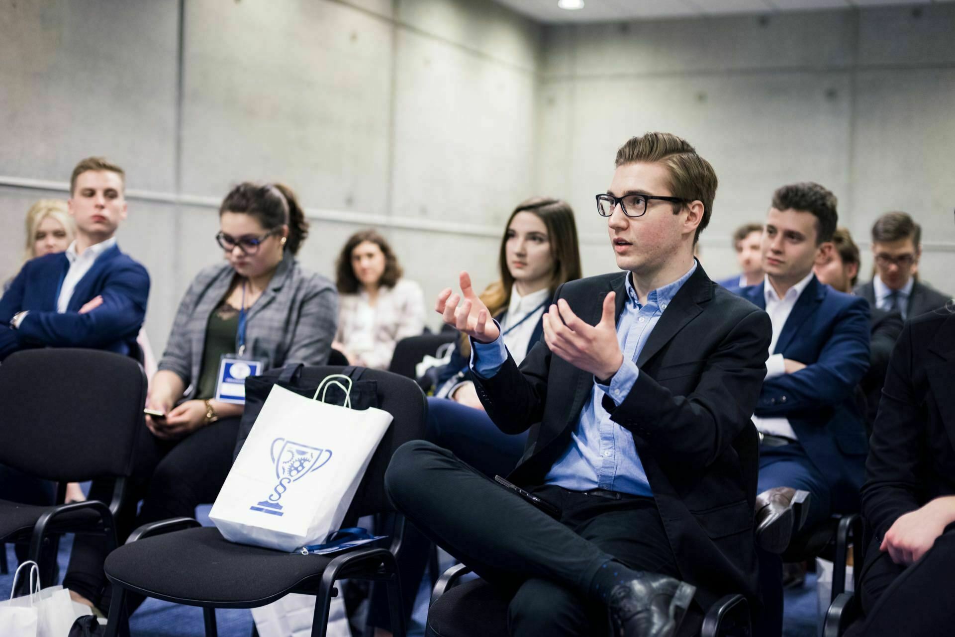 A group of people sitting in a conference room, with a man in the foreground gesturing as he speaks. Some participants have notebooks and bags, and one person is looking at his phone. The room has a modern, minimalist decor with gray walls and a blue carpet - perfect for event photography.  