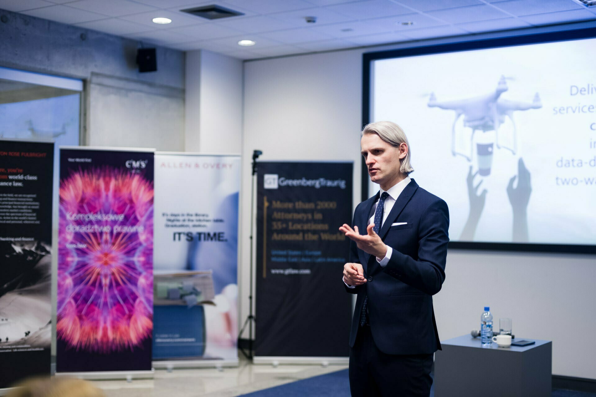 A man in a suit addresses the audience, standing in front of a projection screen displaying a drone image. Behind him, colorful banners with various company logos enliven the professional conference room, making it ideal for any event photo shoot or event photography. 