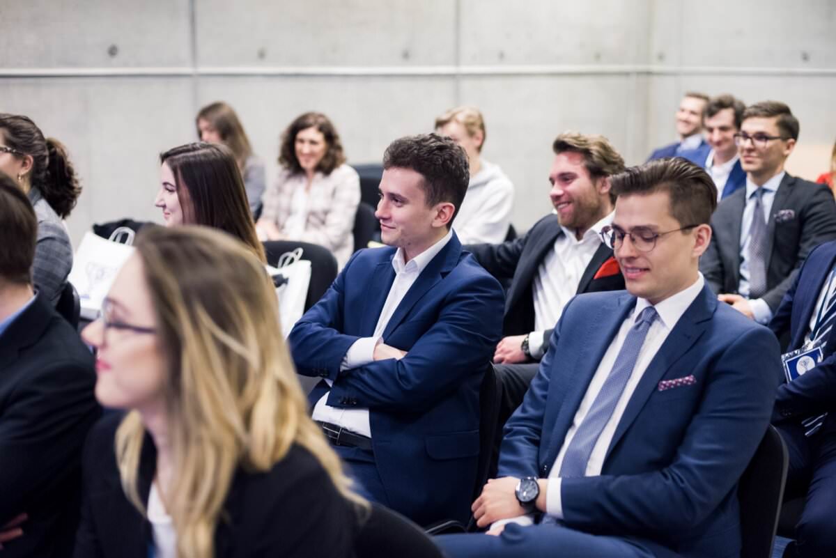 Eine Gruppe von Menschen in formeller Kleidung sitzt in Reihen und hört aufmerksam einer Präsentation oder einem Redner zu. Sie sehen engagiert aus und einige lächeln. Die Szenerie deutet auf eine professionelle oder lehrreiche Veranstaltung hin, die in einem modernen, gut beleuchteten Raum stattfindet, ideal für die Eventfotografie durch den Eventfotografen Warschau.