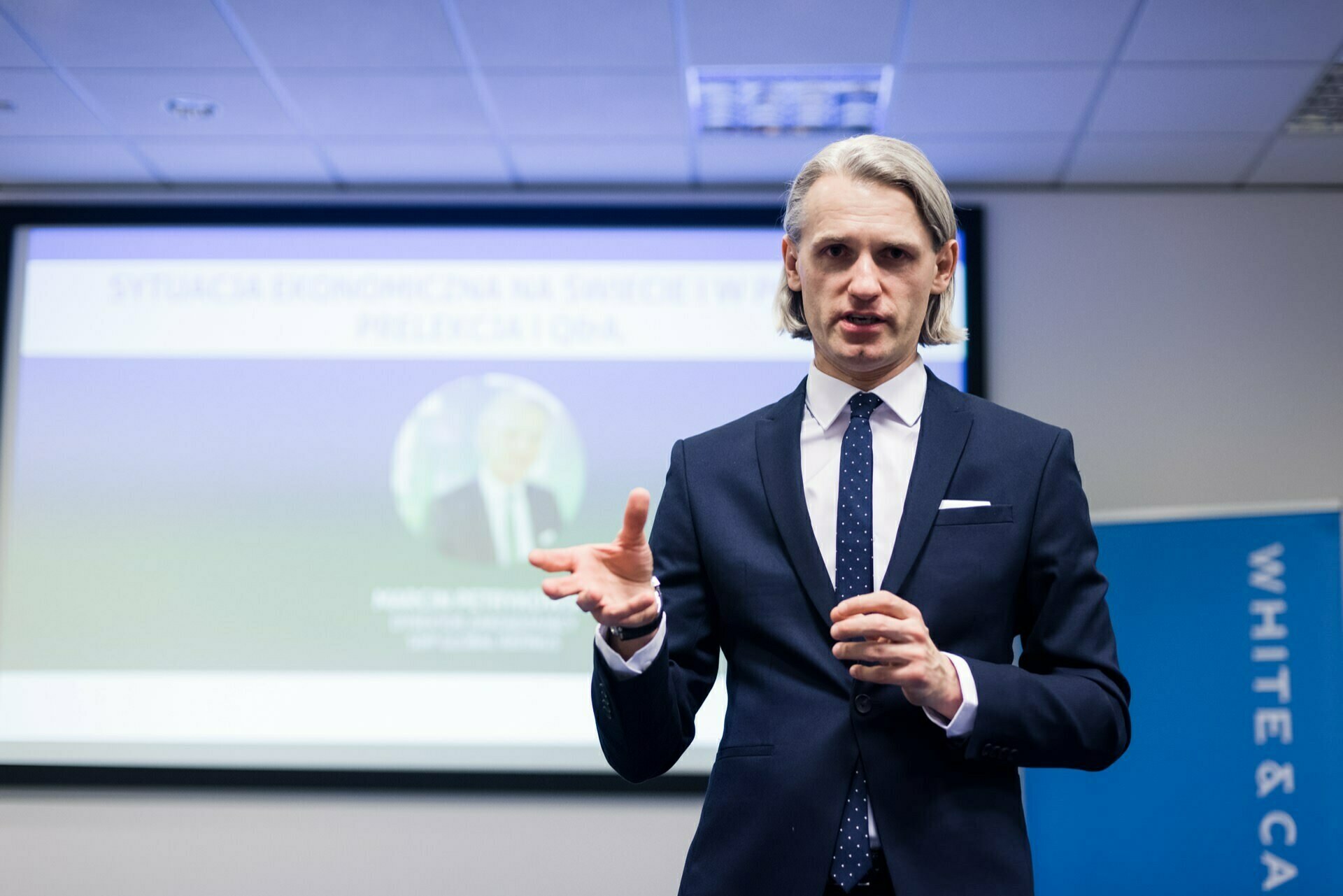 A man in a suit and tie is giving a presentation in a conference room. He gestures with one hand. Behind him, a blurry slide is visible on the screen. In the background is a section of a banner with partially visible text, captured during a photo recap of the events.   
