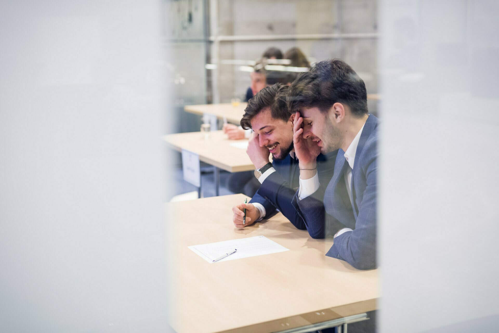 Two men in business attire sit at a table, working closely over a piece of paper. They are smiling and appear to be engaged in discussion. The photo, taken through a glass partition, exudes sincerity and spontaneity, perfect for presentation in a professional event photo essay.  