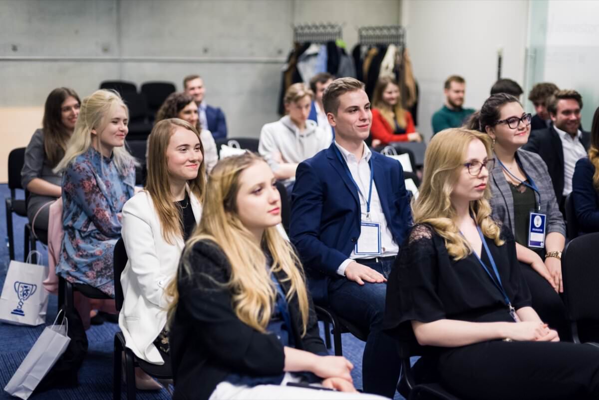 Eine Gruppe junger Erwachsener sitzt in einem Konferenzraum und hört aufmerksam einer Präsentation zu. Sie sind halb-formal gekleidet und tragen Namensschilder. Sie hören aufmerksam zu, während das moderne Interieur mit einer Garderobe und ein paar Stühlen im Hintergrund die Kulisse für diese Eventfotografie bildet.