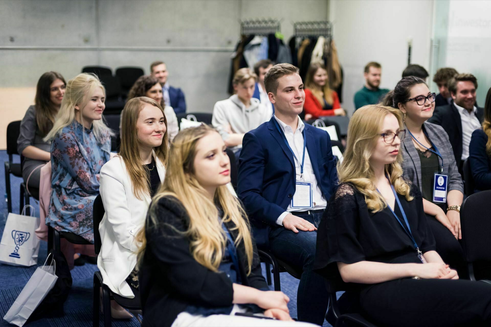 A group of young adults sitting in a conference room and listening intently to a presentation. Dressed in semi-formal attire and wearing name badges, they listen intently as the modern interior, complete with a coat rack and a few chairs in the background, provides the setting for this event photography. 