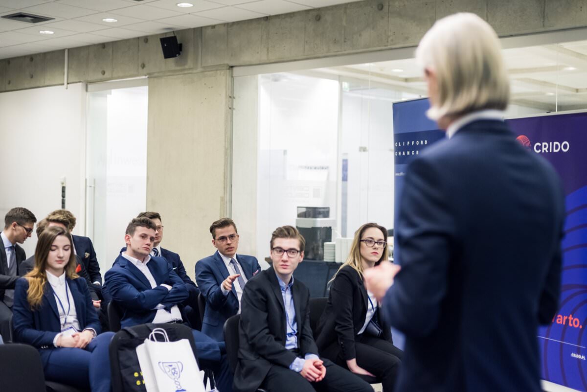 Eine Gruppe von Personen in Geschäftskleidung sitzt und hört aufmerksam einem stehenden Redner bei einer formellen Veranstaltung oder einem Meeting in einem modernen Konferenzraum zu. Hinter dem Redner ist ein Banner mit dem Wort 'CRIDO' und einem blauen Design zu sehen, ideal für alle Eventfotografie-Bedürfnisse.