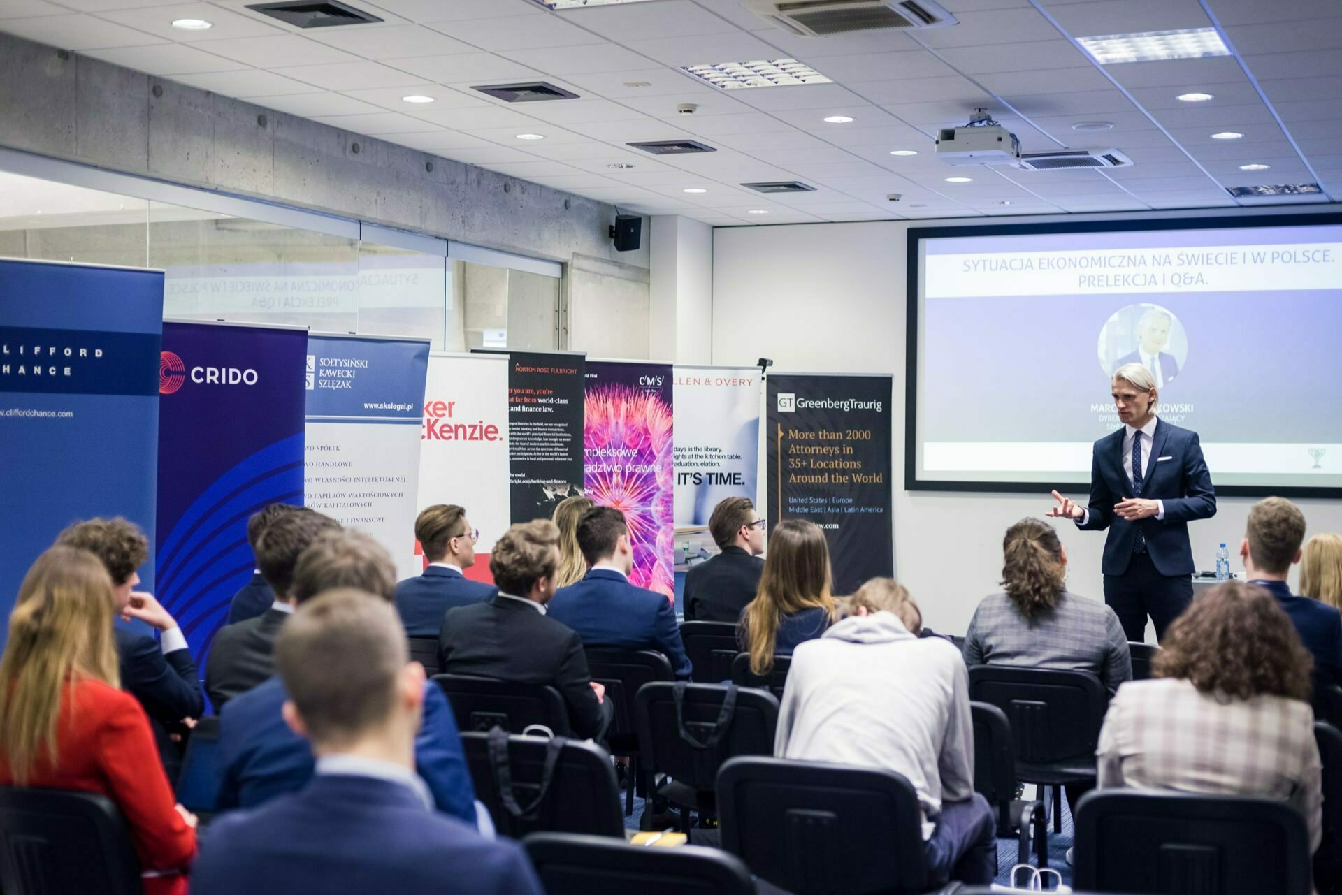 A man in a suit is giving a presentation before an audience seated in rows in a conference room. Behind him, a large screen displays the slides, alongside various company banners and posters arranged along the wall. Photo coverage of the event captures this professional meeting, where participants are dressed in business attire.  