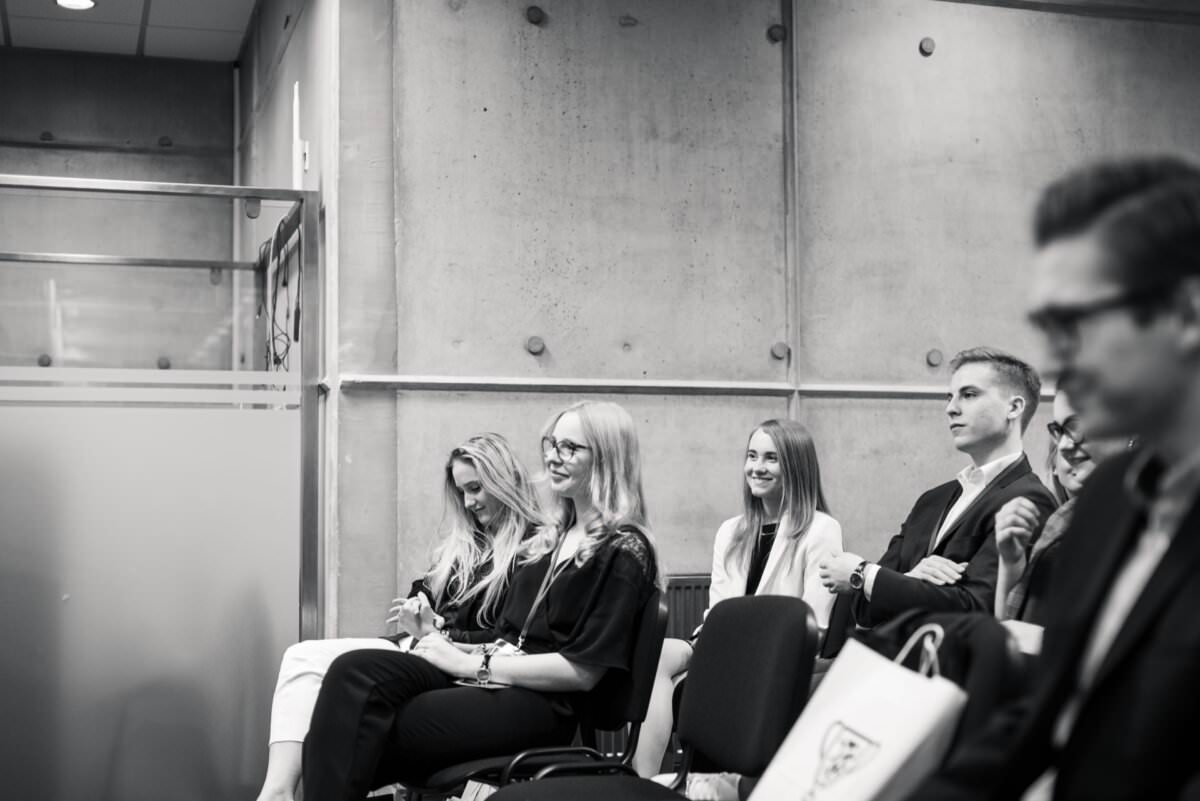 Das schwarz-weiße Veranstaltungsfoto zeigt eine Gruppe von Menschen, die in einem Raum sitzen und aufmerksam etwas außerhalb des Rahmens beobachten. Der Schauplatz ähnelt einer Konferenz oder einem Seminar, mit einer Betonwand im Hintergrund und einer Glaswand auf der linken Seite. Diese Eventfotografie bietet einen Einblick in einen immersiven Moment.