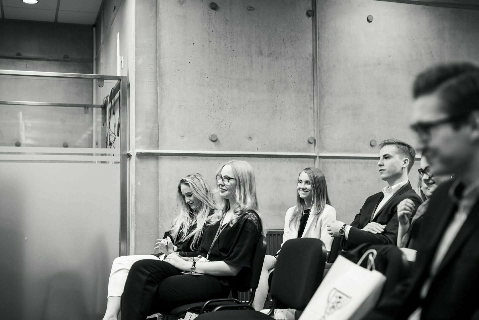 The black-and-white event photograph shows a group of people sitting indoors and carefully observing something outside the frame. The venue resembles a conference or seminar, with a concrete wall in the background and a glass partition to the left. This event photo gives a glimpse of an immersive moment.  