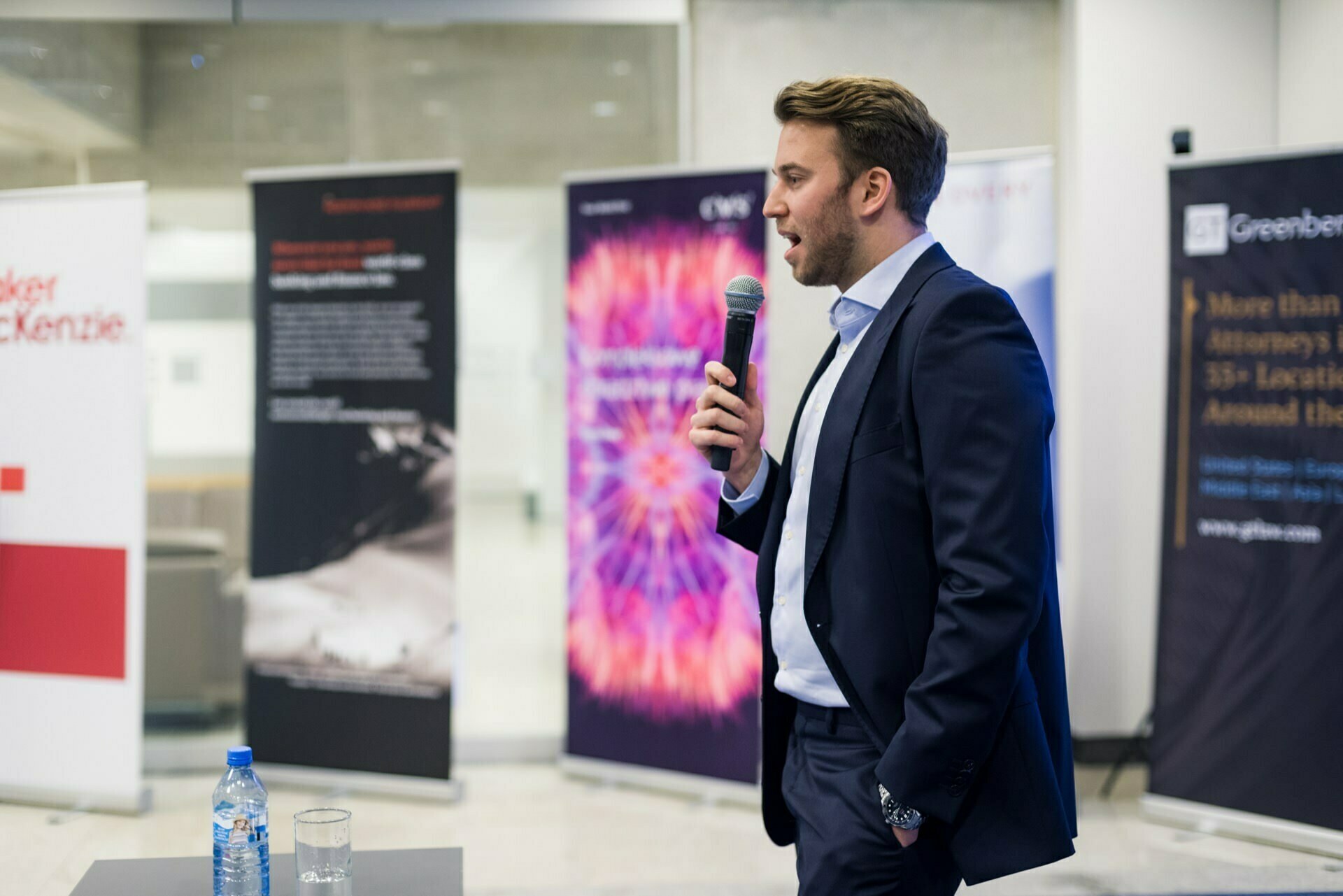 A man in a suit is speaking into a handheld microphone at an event being held indoors. He is standing in front of several banners and a table with a bottle of water and other items. The background is a modern room with blurred people and objects, capturing the essence of professional event photography.  