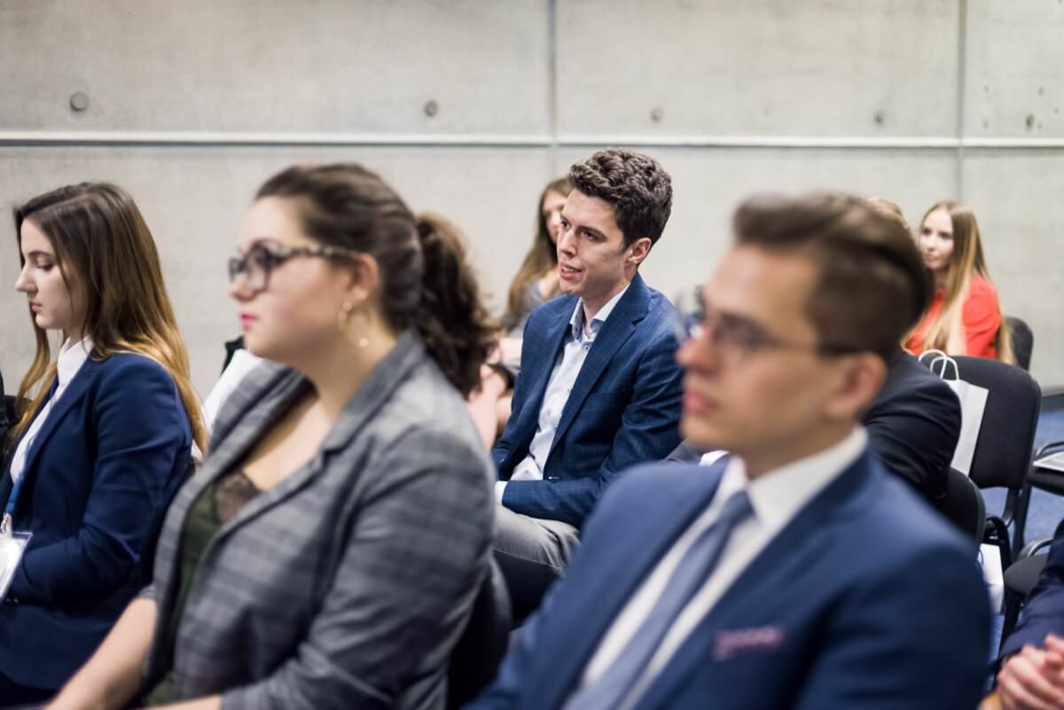In Reihen sitzende Menschen, die an einem Seminar oder einer Konferenz in einem modernen, minimalistischen Raum teilnehmen. Unter den Teilnehmern stellt der Mann in der Mitte eine Frage, während die anderen aufmerksam zuhören. Alle sind in Business-Kleidung gekleidet, die der Event-Fotograf Warschau perfekt eingefangen hat, um eine perfekte Fotoreportage der Ereignisse zu erstellen.