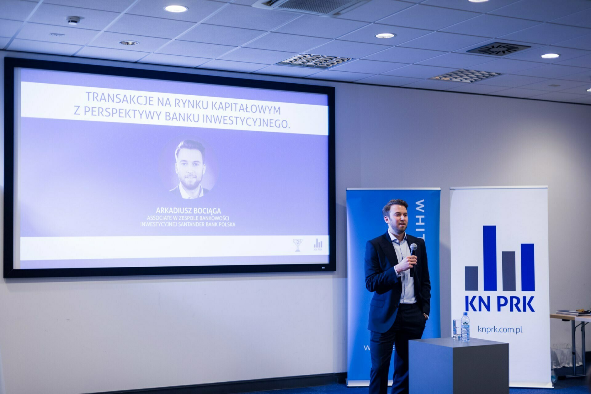 A man is giving a presentation in a modern conference room. An event photography slide displayed on the screen shows the speaker with text in Polish. The room is well-lit in light blue, and there is a podium with a microphone near the speaker.  