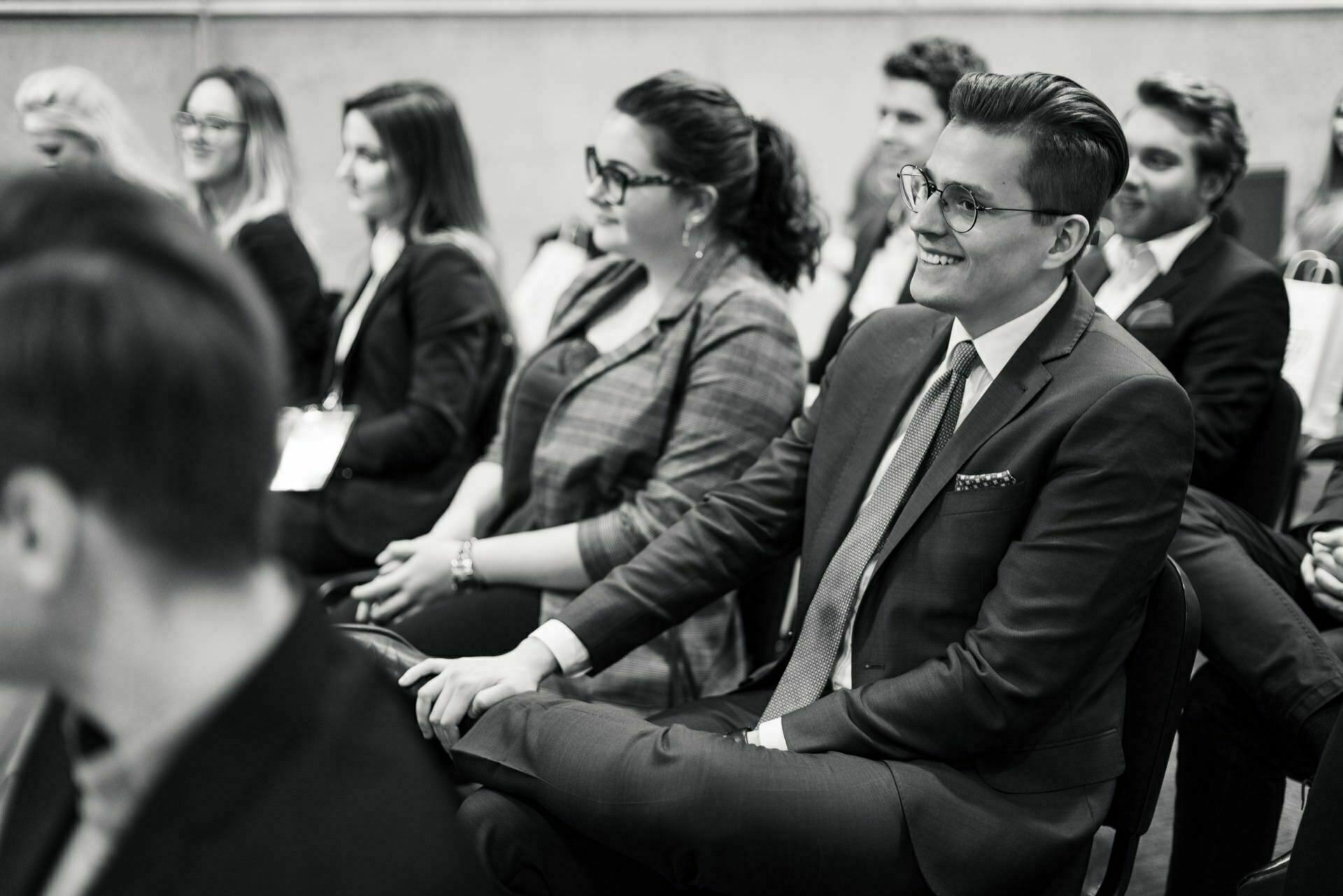 The black-and-white image depicts people sitting in the audience, with an emphasis on a smiling man in a suit and glasses in the foreground. Others around him look forward attentively. The ambiance creates a formal and professional atmosphere, perfectly capturing the moments of event photography.  