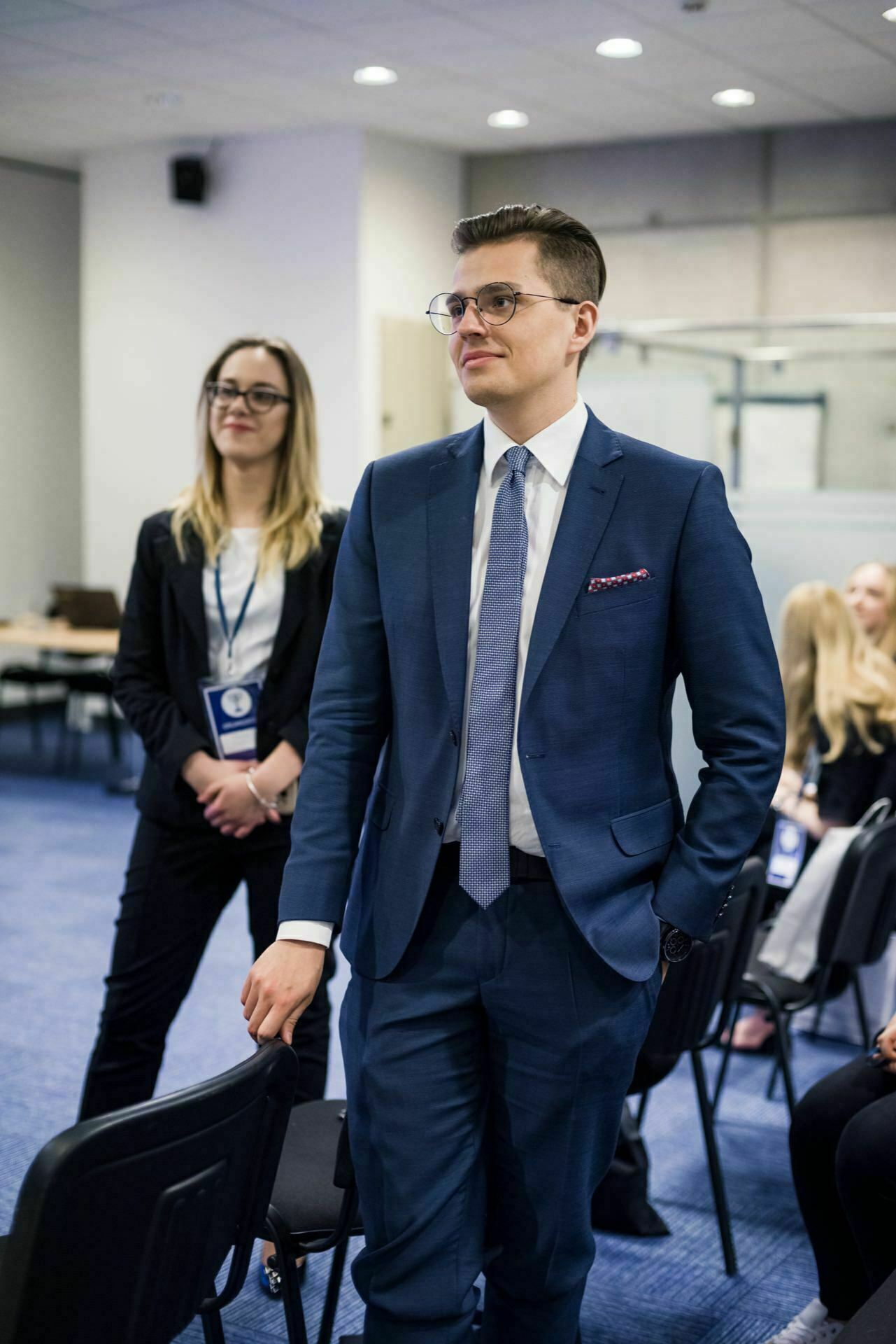 A man in a blue suit stands with his hands in his pockets and looks to the side. He has short hair and wears glasses. Behind him, a woman in a black jacket and glasses smiles. The scenery is reminiscent of an office or conference room with blurry attendees in the background, which is beautifully captured by event photography.   