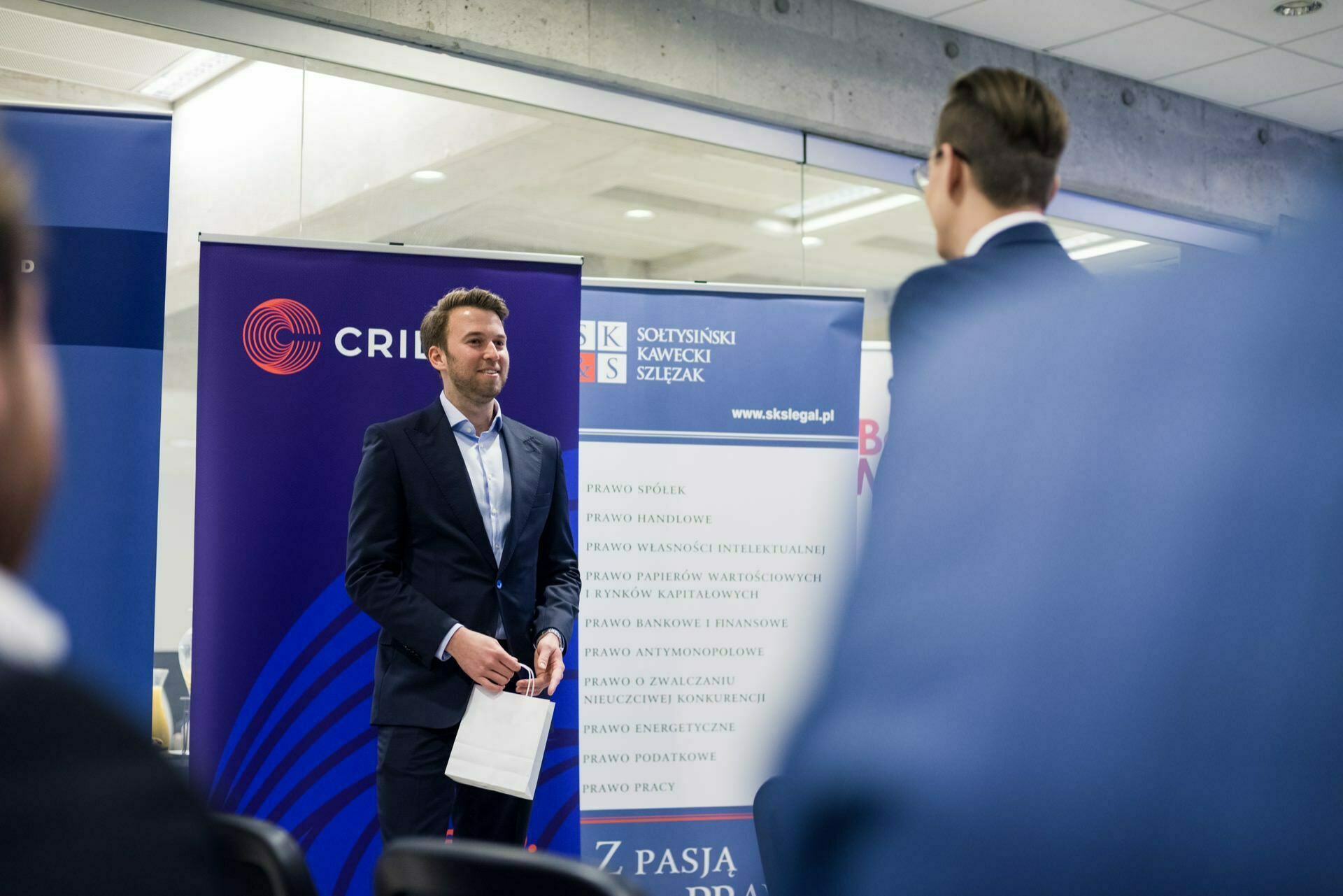 A man in a suit is standing in front of the banners and holding a small gift bag. He appears to be speaking or performing in front of a small audience. The banners behind him display various logos and texts related to legal and business topics. In the foreground, another person can be seen capturing a photo moment of the events.   