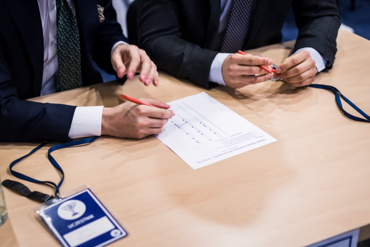 Zwei Personen in Anzügen sitzen an einem Tisch, halten Stifte in der Hand und füllen ein Formular aus. Ein Schlüsselband mit einem Namensschild und einem Ausweis liegt auf dem Tisch und unterstreicht ihre Professionalität. Diese Szene fängt die Essenz der Eventfotografie in Warschau ein und unterstreicht die Liebe zum Detail in jeder Eventfotoreportage.