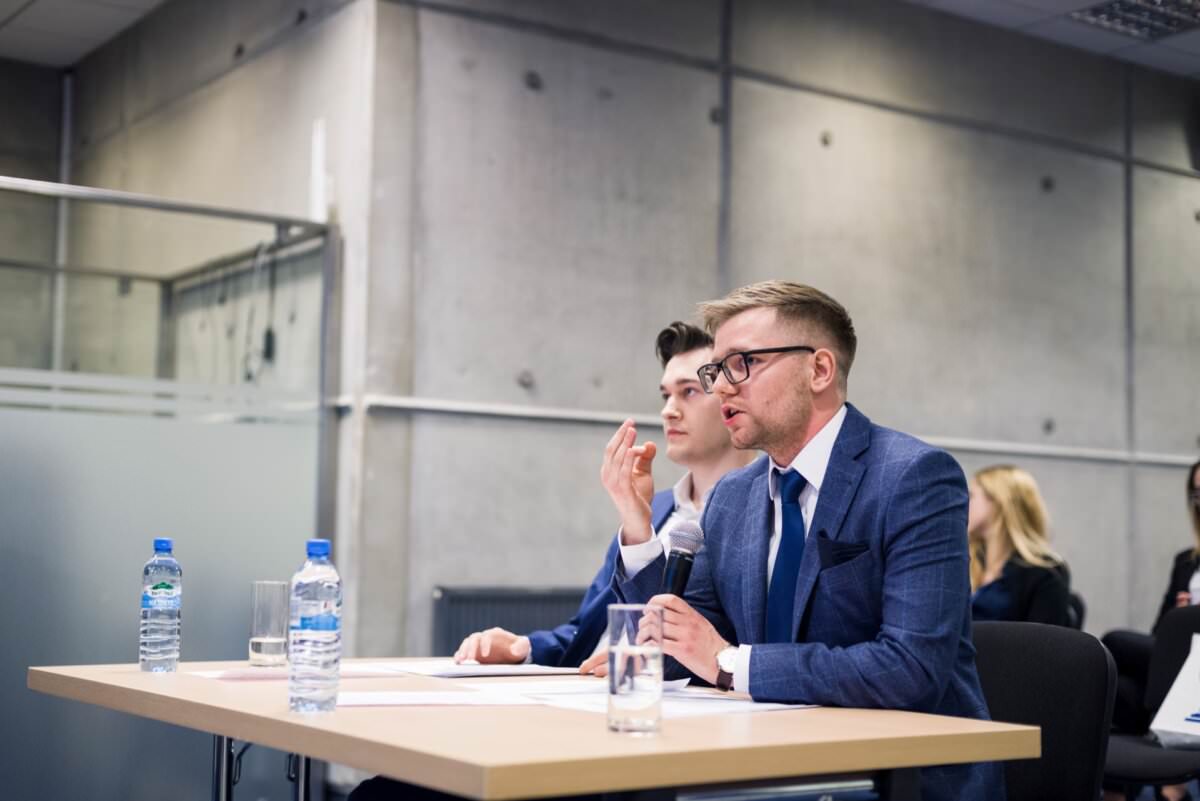 Zwei Männer in Anzügen sitzen an einem Tisch mit Mikrofonen während eines formellen Treffens oder einer Podiumsdiskussion in einem modernen Konferenzraum. Ein Mann spricht, der andere hört aufmerksam zu. Auf dem Tisch stehen Wasserflaschen und Dokumente - ein perfektes Beispiel für Eventfotografie, die die Essenz solcher Veranstaltungen einfangen kann.