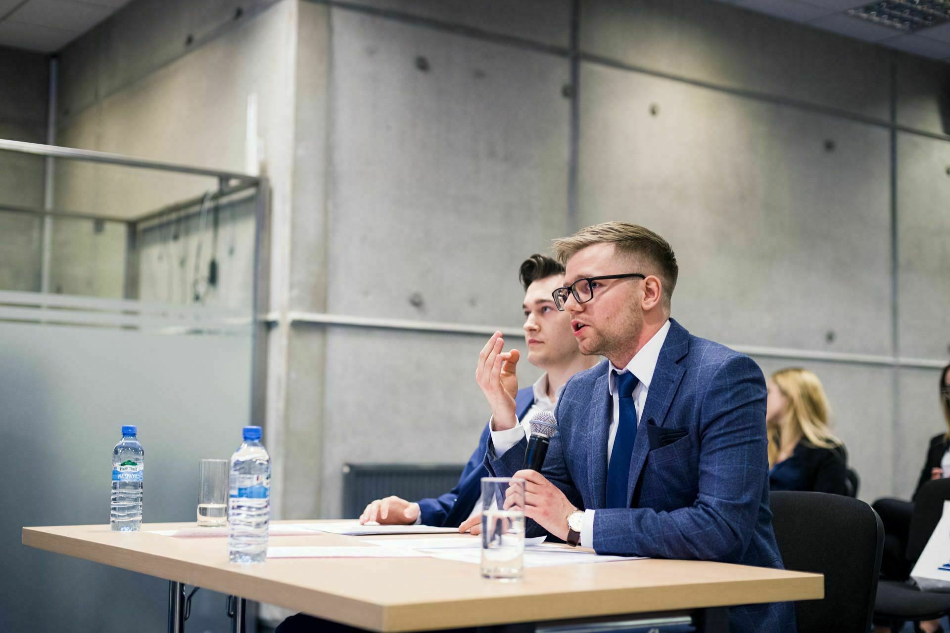 Two men in suits sit at a table with microphones during a formal meeting or panel discussion in a modern conference room. One man speaks, the other listens intently. Water bottles and documents stand on the table - a perfect example of event photography that can capture the essence of such events.  