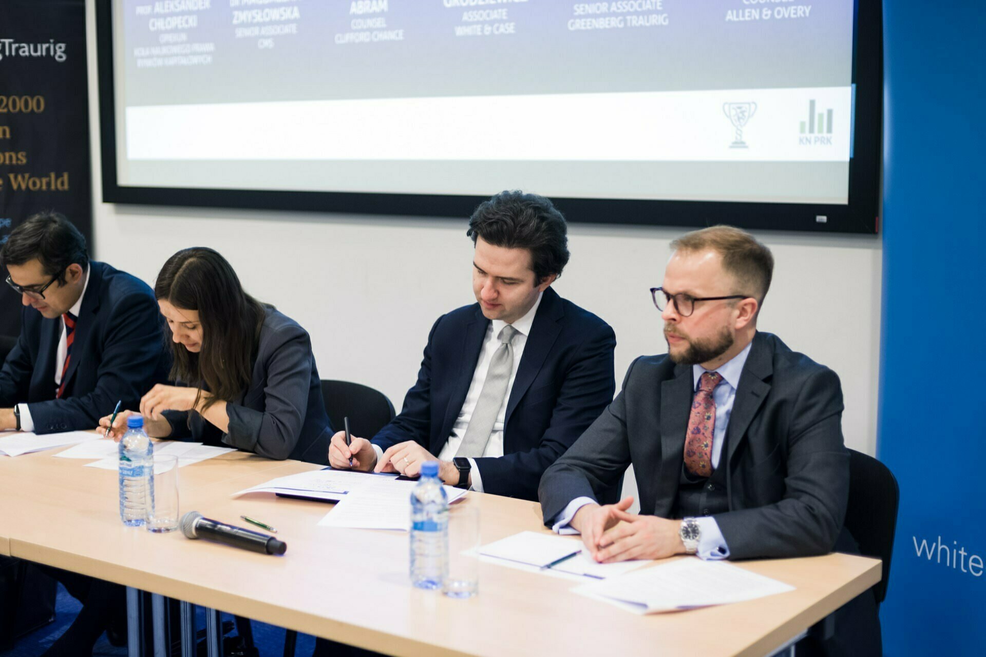 Four professionally dressed people sit at a table in front of which are papers and water bottles. They look focused, writing notes. Behind them is a screen with text, and to the right is a partially visible blue banner - a snapshot perfect for event photography.  