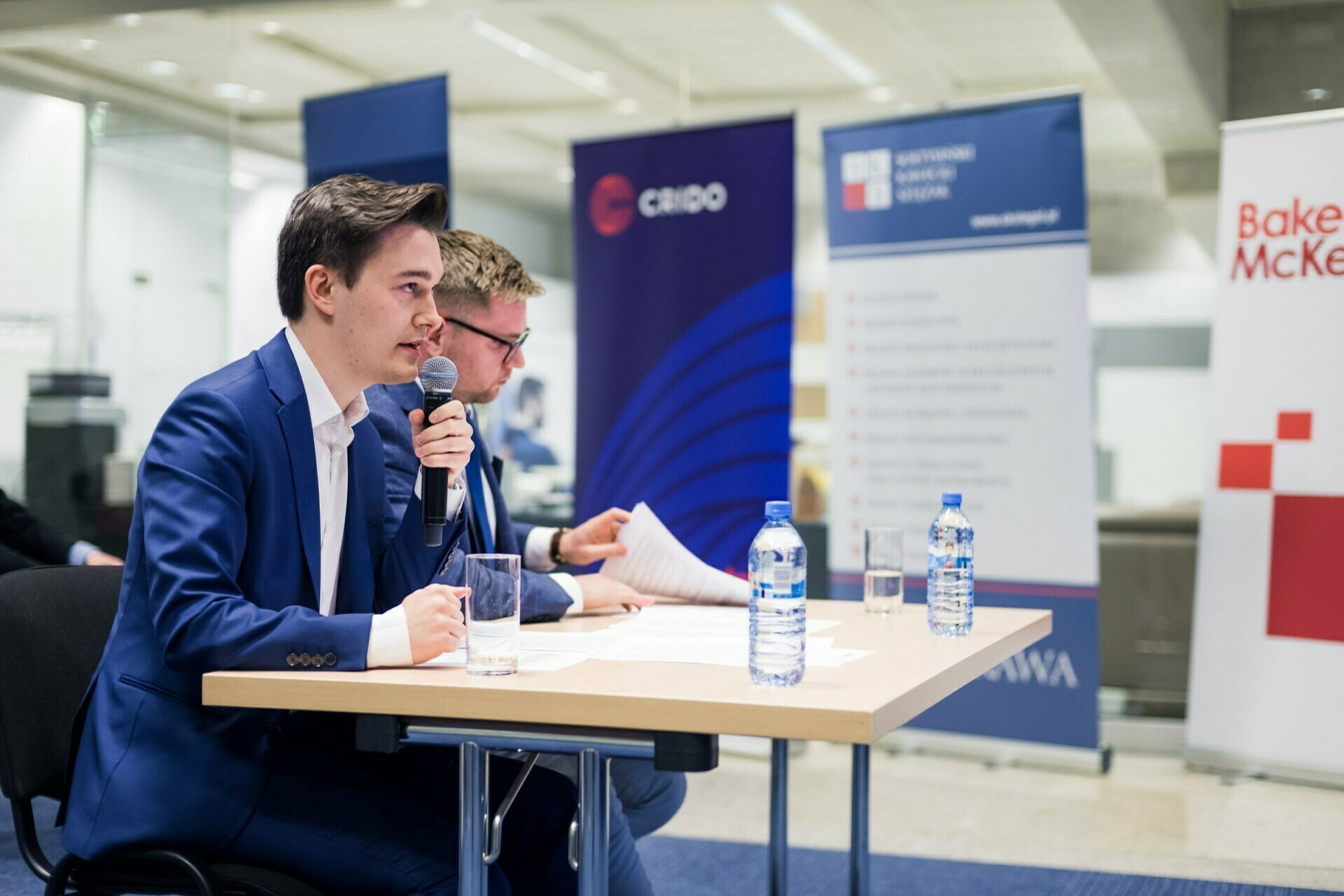 Two men are sitting at a table in a conference room. The man on the left, wearing a blue suit, is speaking into a microphone, holding a pen. The man on the right, also wearing a suit, appears to be reading. On the table lie bottles of water and papers. This scene is perfect for any event photographer Warsaw who wants to capture dynamic moments.    