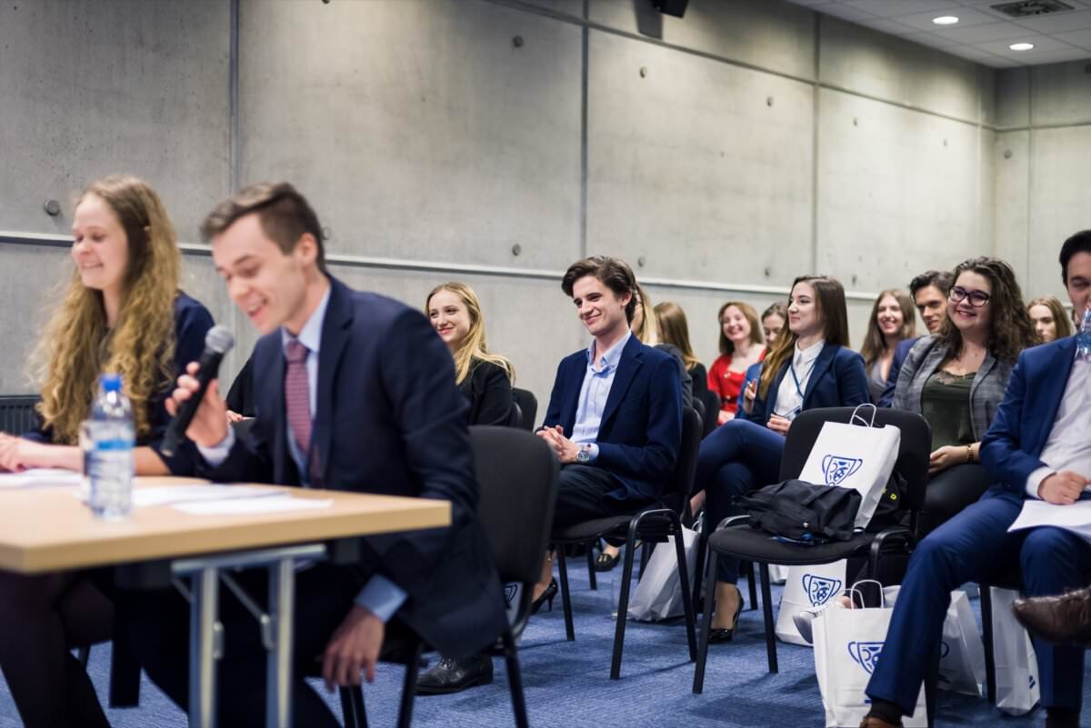 Eine Gruppe von Personen in formeller Kleidung sitzt in einem Raum und hört aufmerksam einer Person zu, die an einem Tisch im Vordergrund in ein Mikrofon spricht. Einige Teilnehmer halten Einkaufstaschen in der Hand, was darauf hindeutet, dass es sich um eine Konferenz oder ein Seminar handelt, das von einem Eventfotografen Warschau festgehalten wird, ideal für Event-Fotojournalismus.