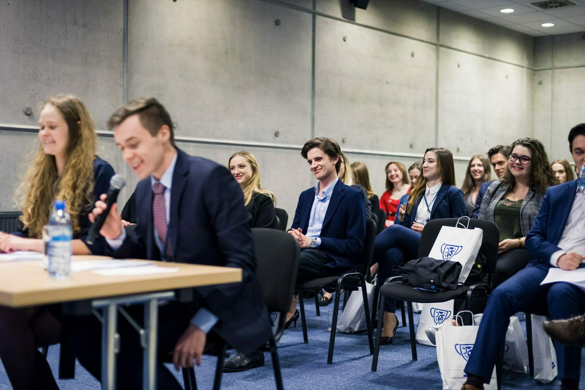 A group of people in formal attire sit in a room and listen intently to a person speaking into a microphone at a table in the foreground. Some of the attendees are holding shopping bags, suggesting that this is a conference or seminar being captured by an event photographer Warsaw, perfect for event photojournalism. 