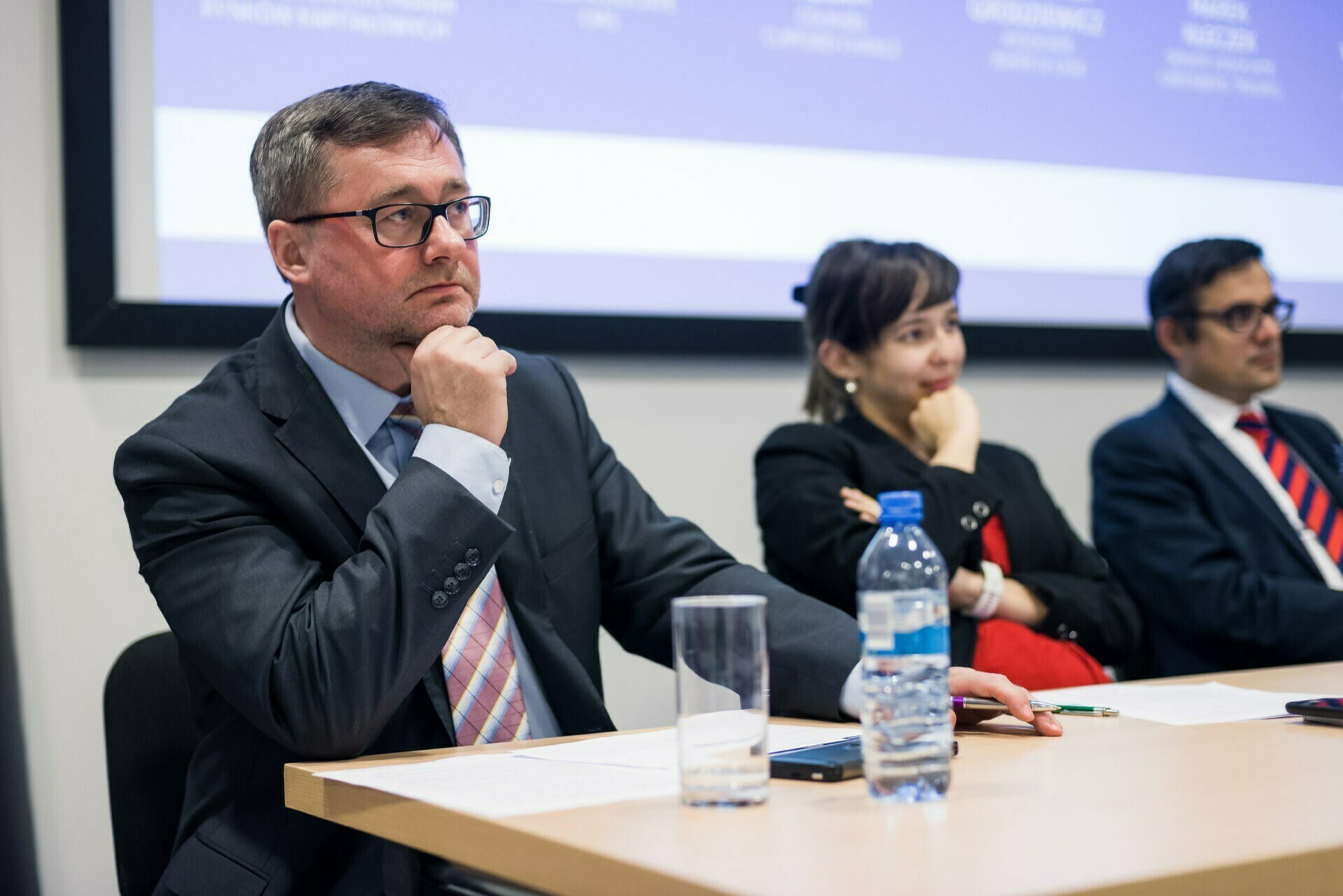 A man in a suit and glasses sits at a conference table with a serious face, resting his chin on his hand. Sitting attentively next to him are a woman and another man, both in business attire. In the background is a presentation screen, and on the table is a bottle of water. This scene captures the essence of event photography at its best.   