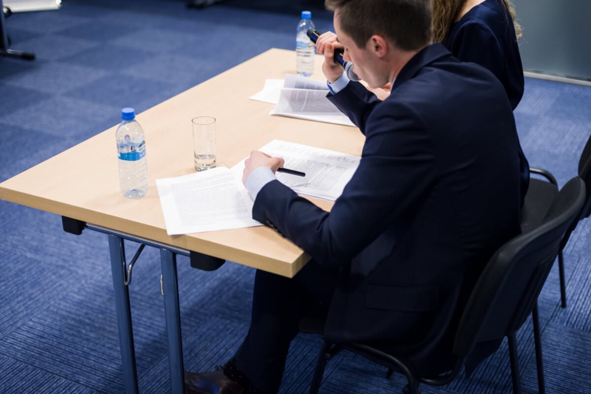 Eine Person im Anzug sitzt an einem Tisch, sieht Dokumente durch und macht sich mit einem Stift Notizen. Eine andere Person ist teilweise neben ihr zu sehen. Wasserflaschen und ein Glas stehen auf einem Tisch in einem Raum mit blauem Teppichboden und fangen einen Moment ein, der sich perfekt für Eventfotografie eignet.