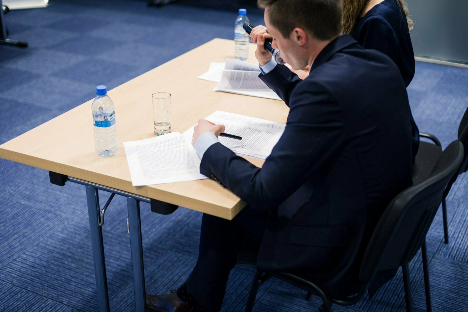 A person in a suit is sitting at a table, reviewing documents and taking notes with a pen. Another person is partially visible next to them. Water bottles and a glass stand on a table in a room with blue carpeting, capturing a moment perfect for event photography.  