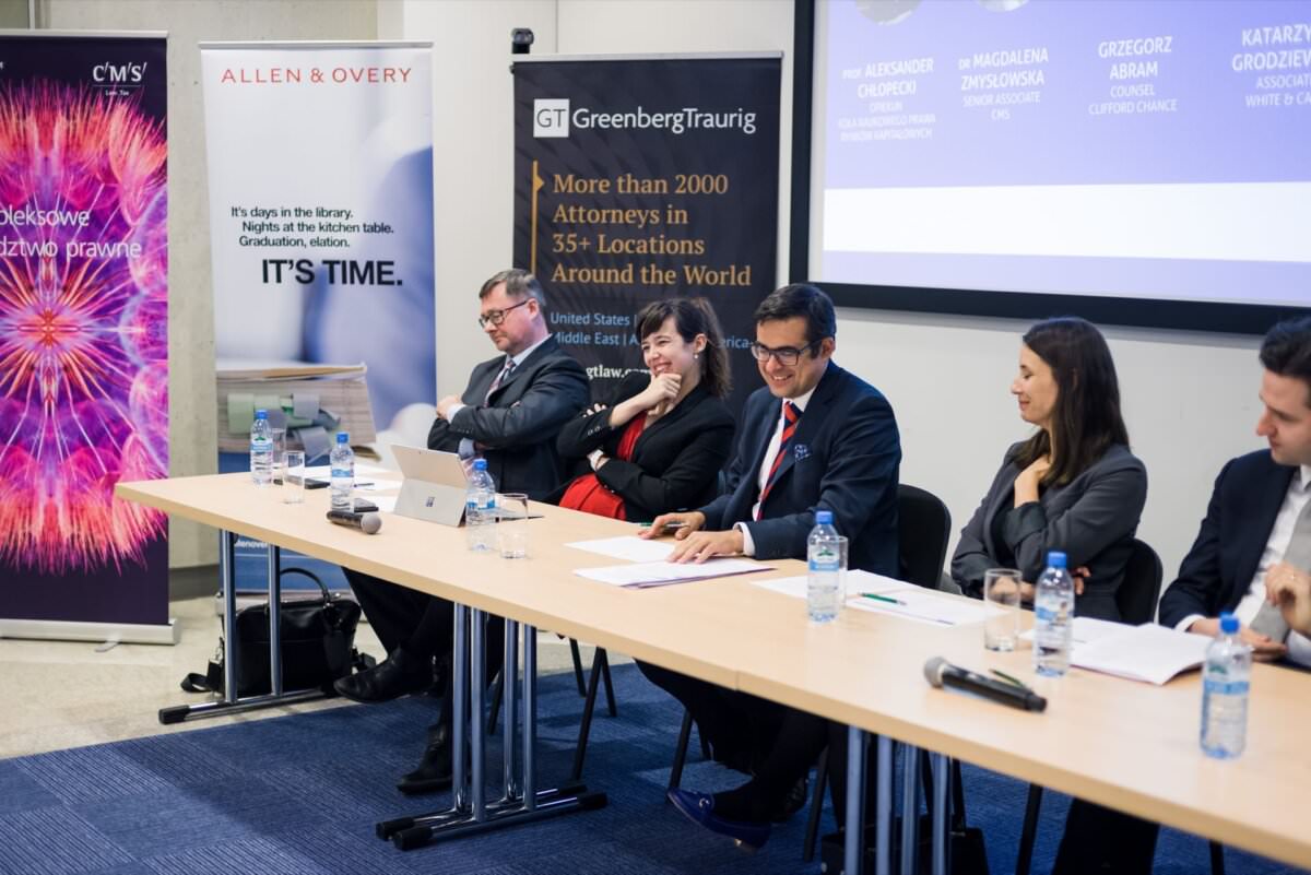Eine Gruppe von fünf Fachleuten sitzt an einem langen Tisch mit Mikrofonen und Wasserflaschen. Hinter ihnen hängen Banner von verschiedenen Anwaltskanzleien, darunter CMS, Allen & Overy und Greenberg Traurig. Die Podiumsteilnehmer erwecken den Eindruck, als seien sie in die Diskussion vertieft, wobei eine Person spricht - die perfekte Bühne für Eventfotografie in Warschau.