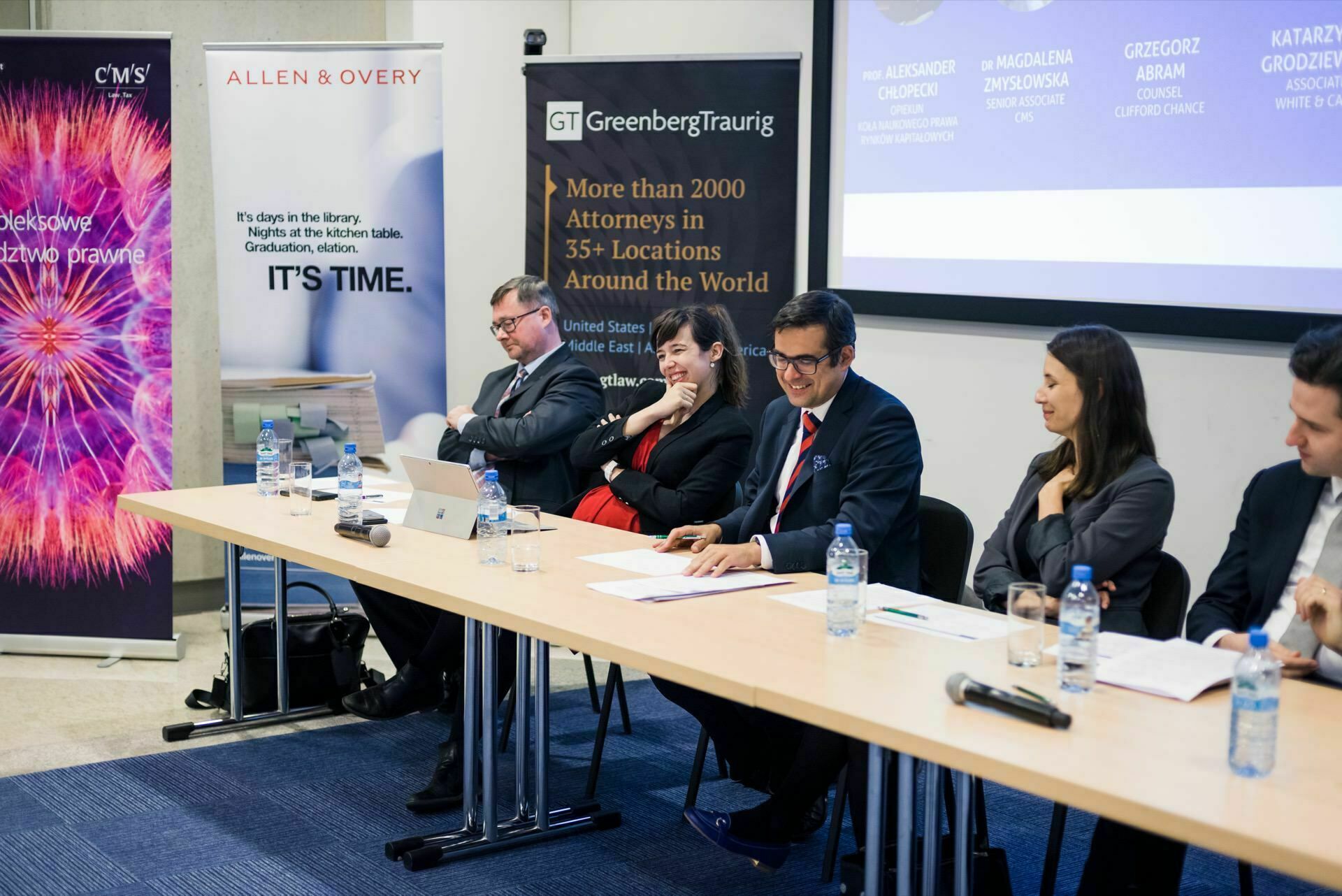 A panel of five professionals sit at a long table with microphones and water bottles. Behind them hang banners of various law firms, including CMS, Allen &amp; Overy and Greenberg Traurig. The panelists give the impression of being involved in the discussion, with one person speaking - a perfect scene for event photography in Warsaw.  