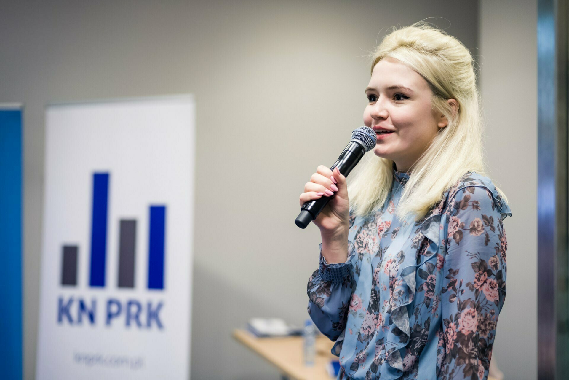 A woman with long blonde hair, wearing a floral blouse, speaks into a microphone at an event taking place indoors. She is standing next to a sign with the logo and text "KN PRK" printed on it. This event photo captures the moment perfectly, with a blurry person and a few blurry objects in the background.  