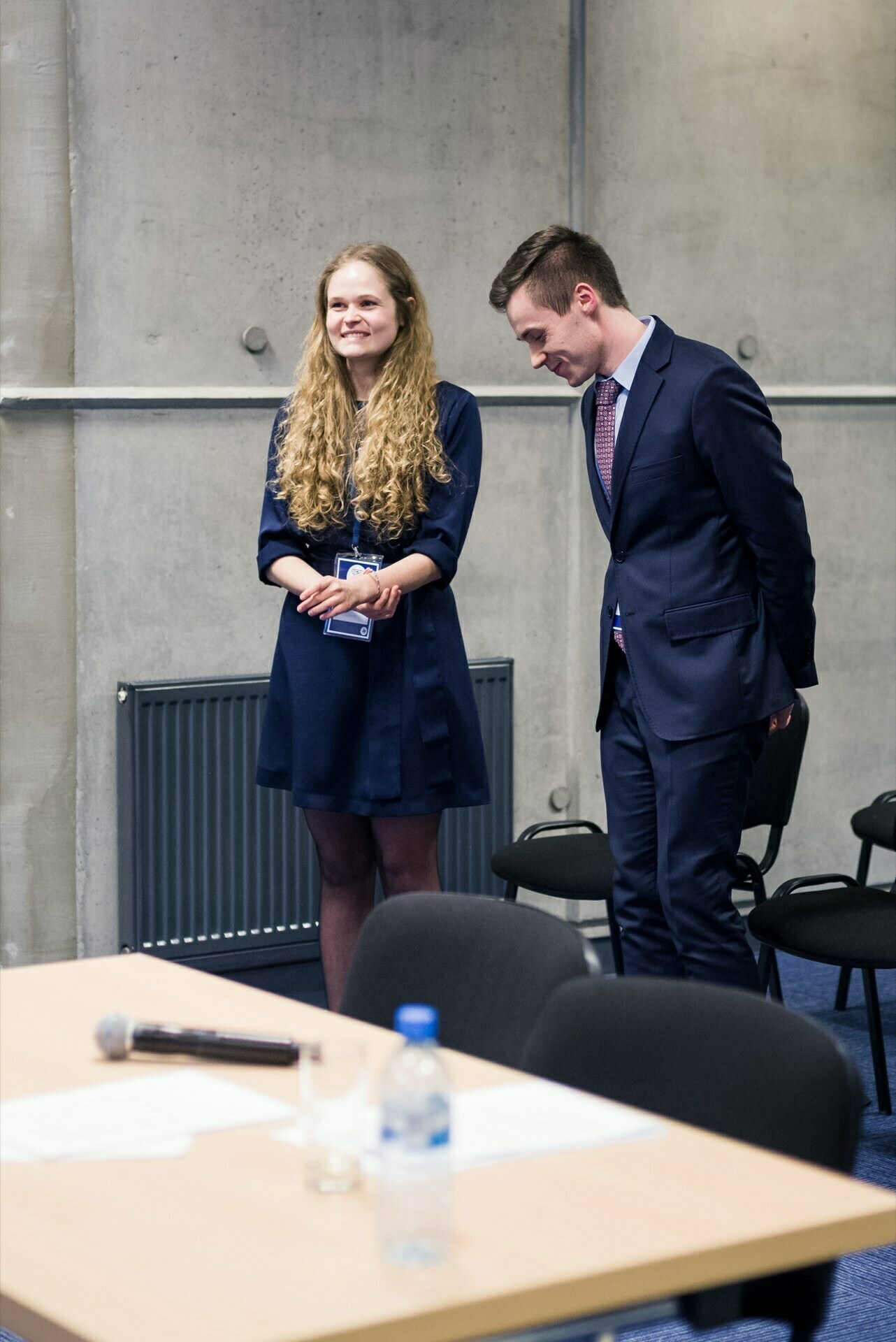 Two people dressed formally stand in a room with a concrete wall and a radiator behind them. The woman on the left has long curly hair and is smiling. The man on the right, dressed in a suit and tie, tilts his head slightly and smiles. In the foreground is a table with documents and a bottle of water - a perfect scene for an event photographer Warsaw capturing photo coverage of events   