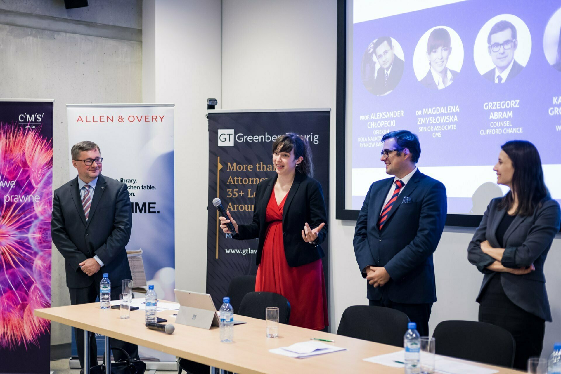 Four people in business attire stand outside a conference room. One woman speaks into a microphone, while the others smile and listen. Behind them are banners and a presentation slide with the names and photos of others - a perfect scene for event photography. In the foreground is a table with bottles of water and papers.   