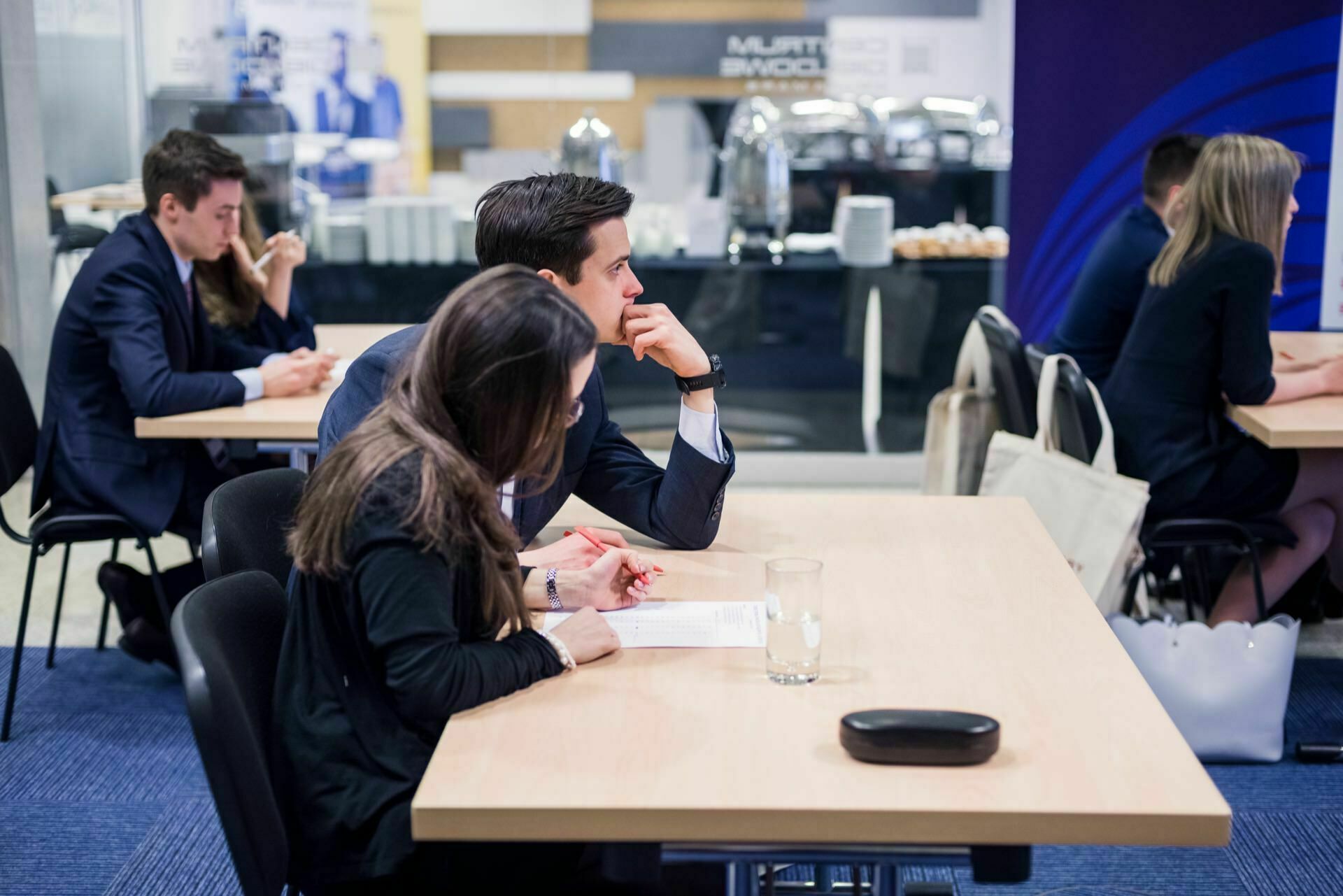 A group of people, mostly in business attire, are sitting at tables, immersed in discussions or working on documents. The venue looks like a professional or networking event, with food and beverage options visible on a buffet table in the background - perfect for event photography. 