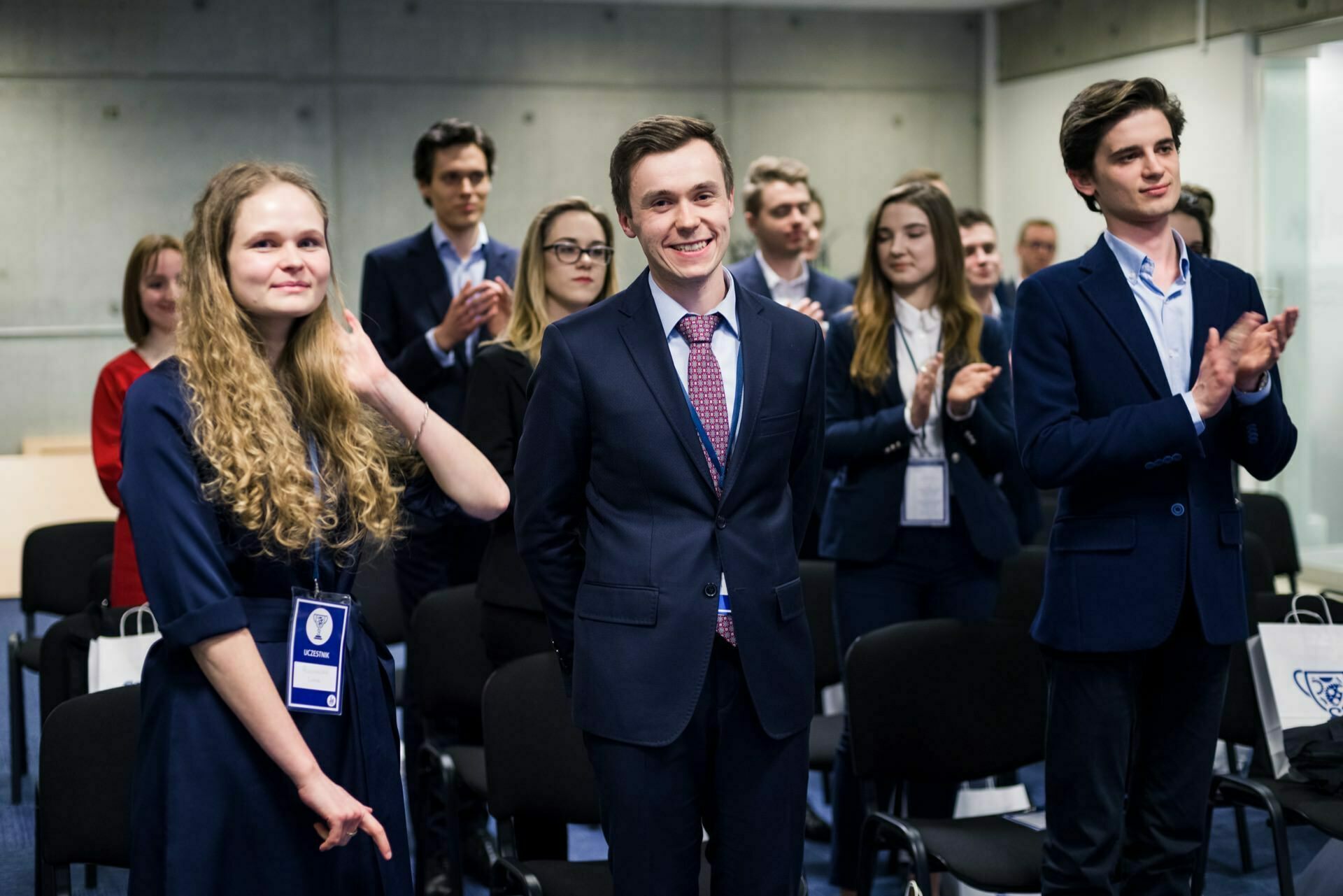 A group of young adults in formal business attire stand and clap in what appears to be a conference or meeting room. The central figure, a man in a suit and tie, smiles. The room has a modern design with white walls and concrete accents - perfect for event photography.  