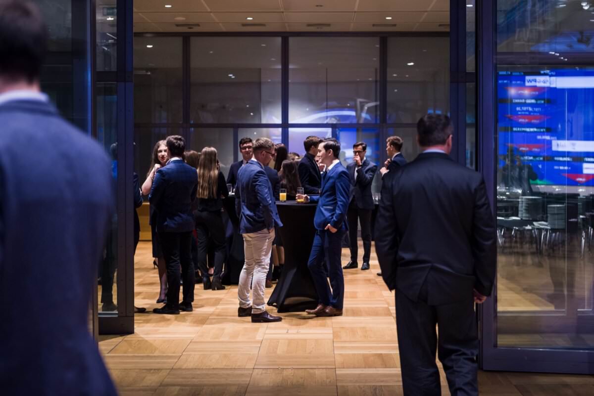 Eine Gruppe förmlich gekleideter Menschen in Anzügen und Kleidern plaudert und trinkt in einem modernen Raum mit großen Glasfenstern und einem Holzboden. Während der Eventfotografie Warschau stehen mehrere Personen in kleinen Gruppen und einige wenige stehen alleine.