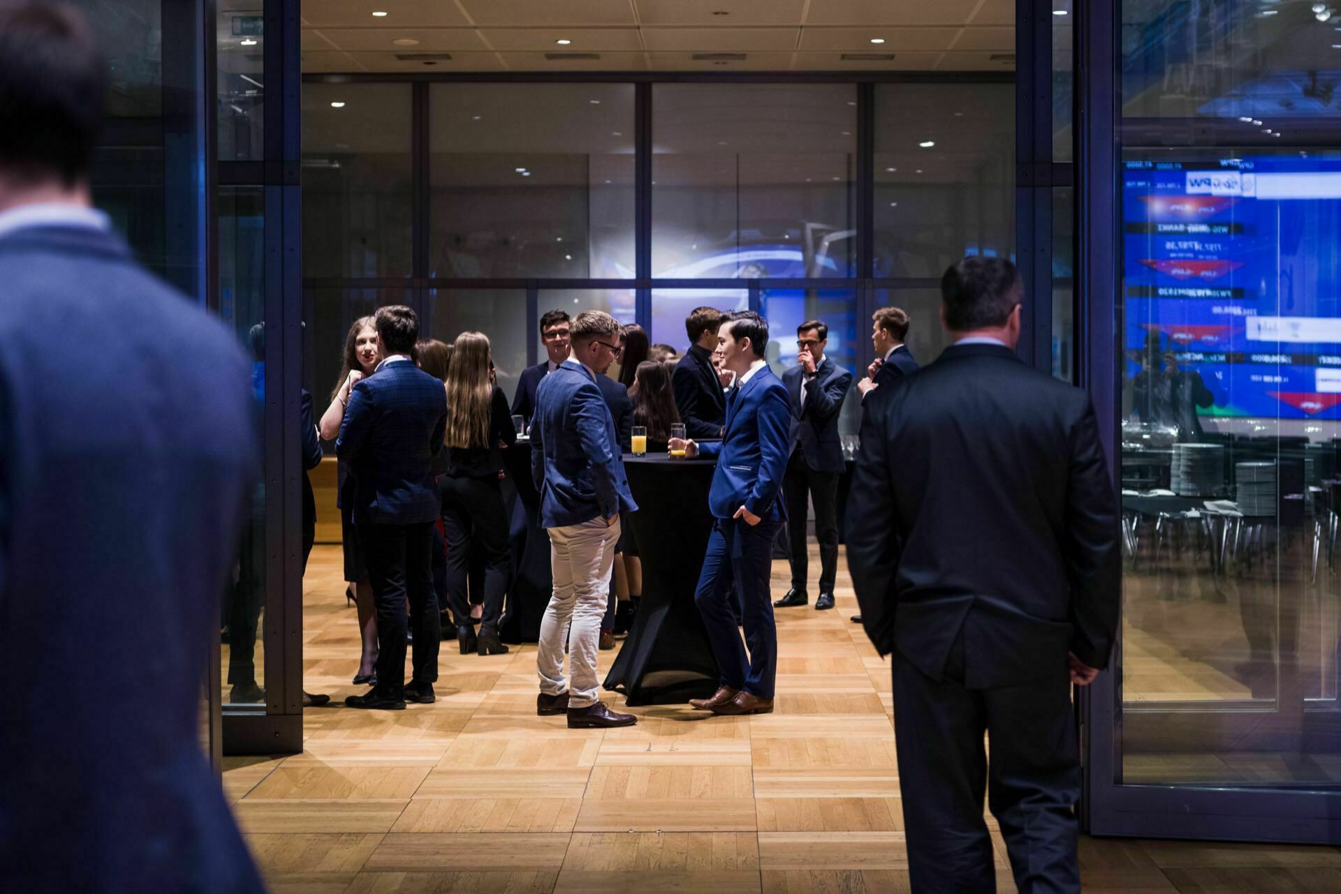 A group of people dressed formally, including in suits and dresses, are chatting and having drinks in a modern room with large glass windows and a wooden floor. During event photography warsaw, several people stand in small groups, while a few stand alone. 