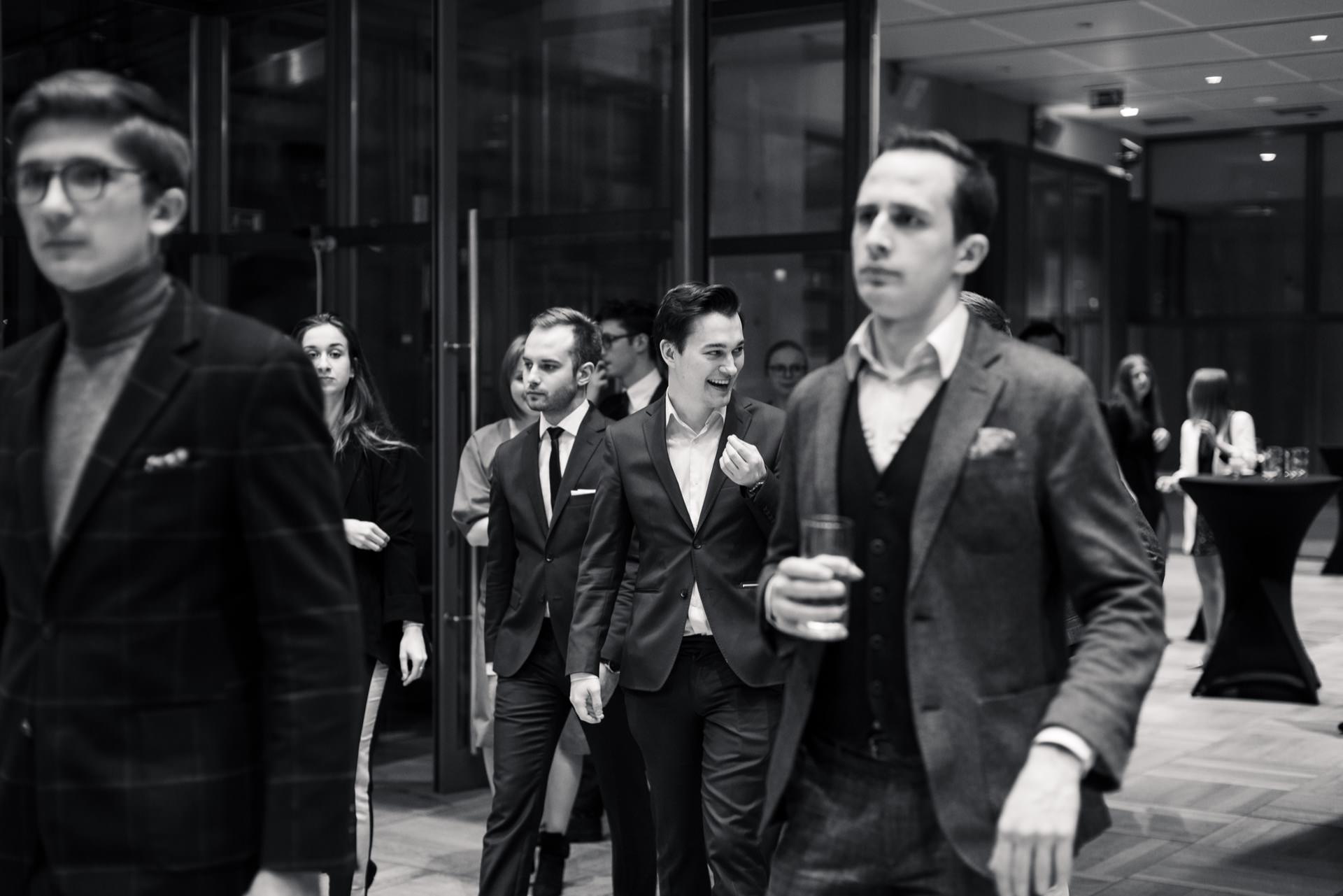 A black and white photo of a group of young adults in formal attire walking through a modern indoor space. Individuals appear to be chatting and smiling while carrying drinks. This event photo captures the lively atmosphere of an elegant social gathering.  