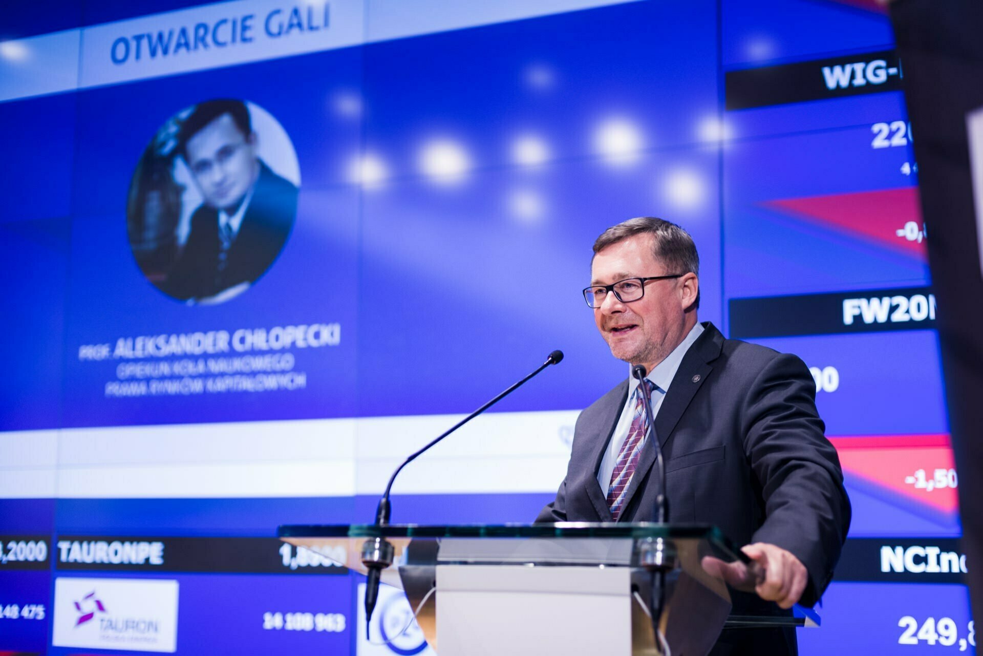 A man in a suit and tie speaks at a podium using a microphone. Behind him, a large screen displays financial information and a photo of another man. The title on the screen reads "GALA OPENING," and there is additional text below that captures the essence of the moment of this event photography in Warsaw.  